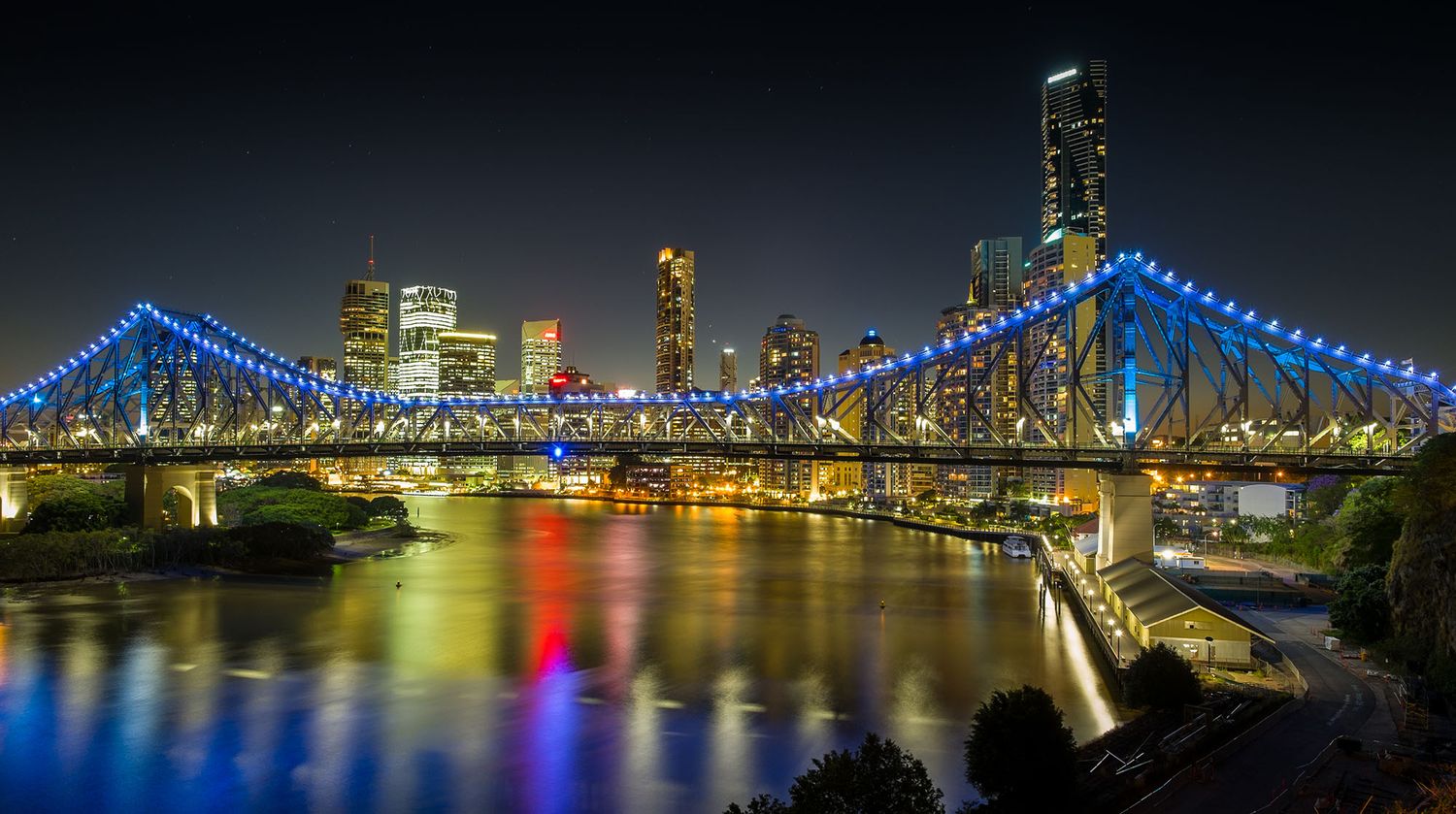 Story Bridge illuminated with blue lights reflecting on Brisbane River at night with city skyline.