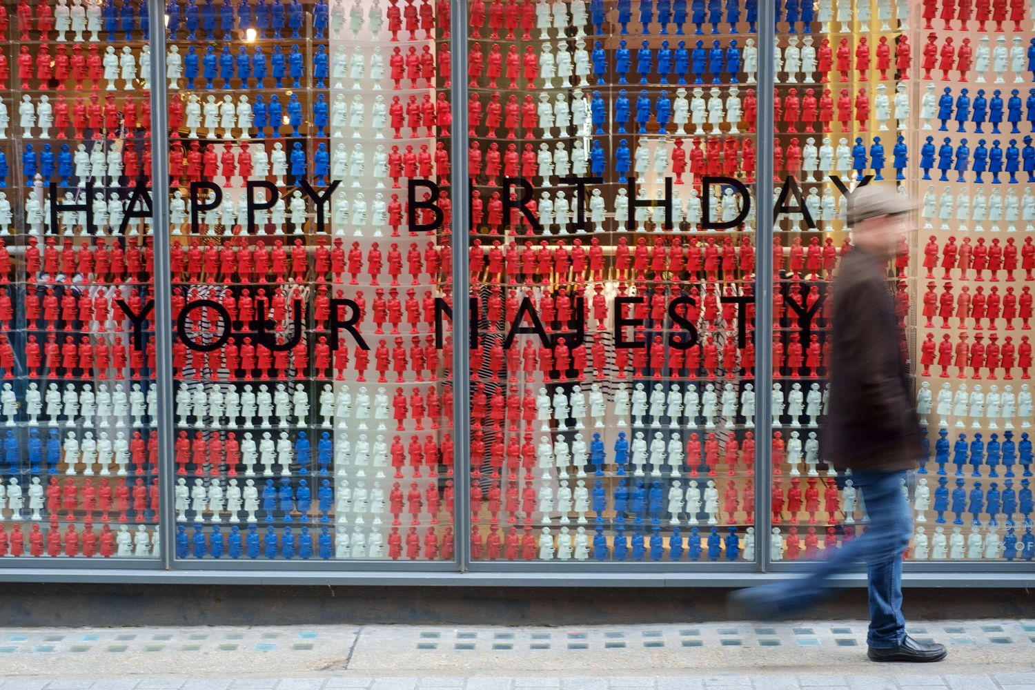 A person walks past a colorful shop window displaying Happy Birthday Your Majesty in bold letters.