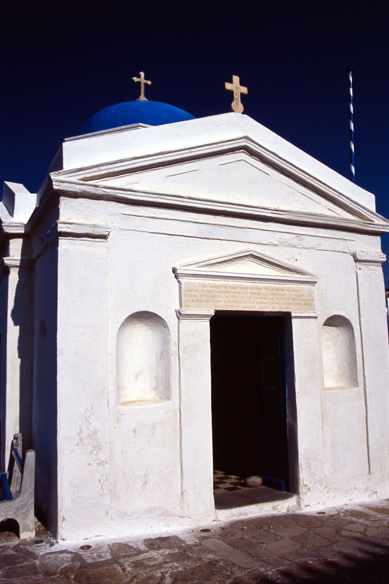 Traditional white Greek Orthodox chapel with blue domed roof and twin crosses under clear blue sky.