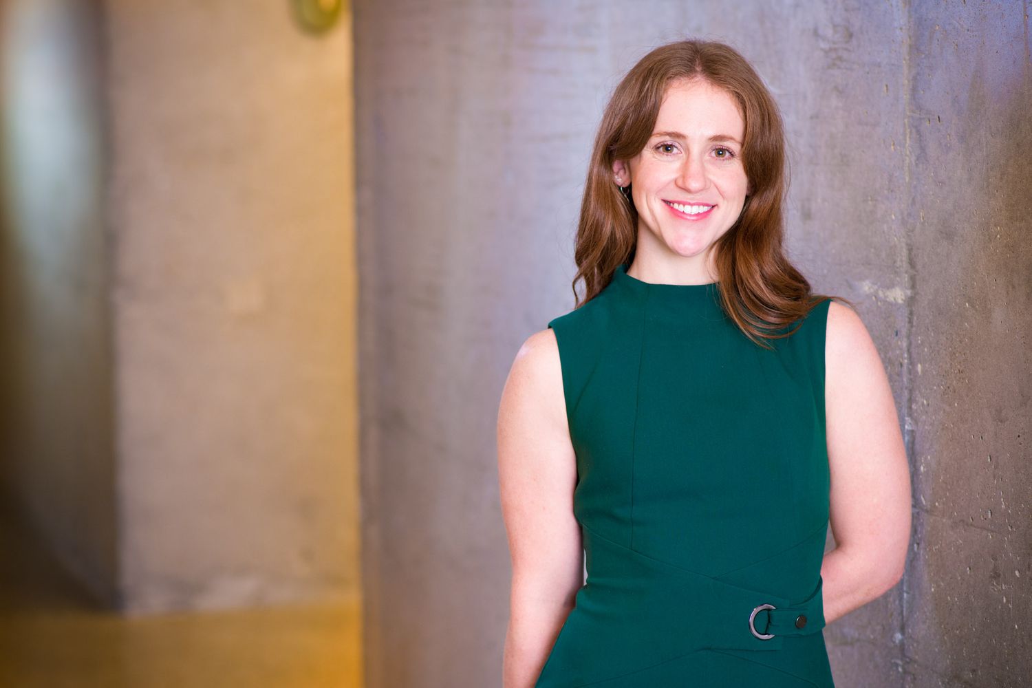 Smiling woman in teal sleeveless dress leaning against concrete pillar in soft lighting.