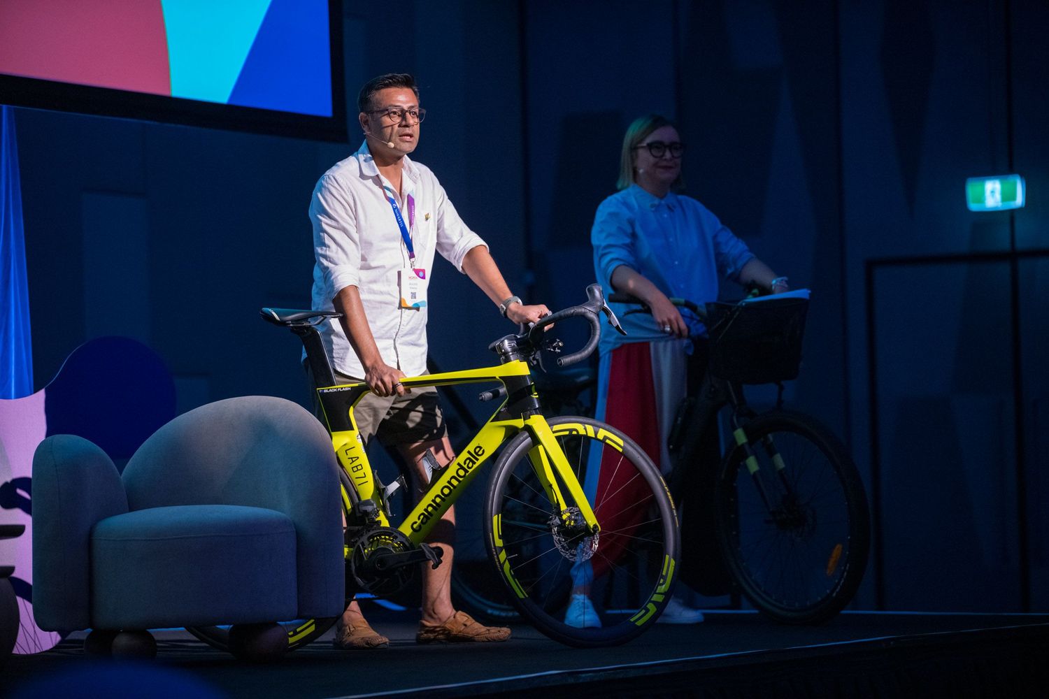 Man in white shirt stands beside a yellow racing bike on a conference stage presentation.