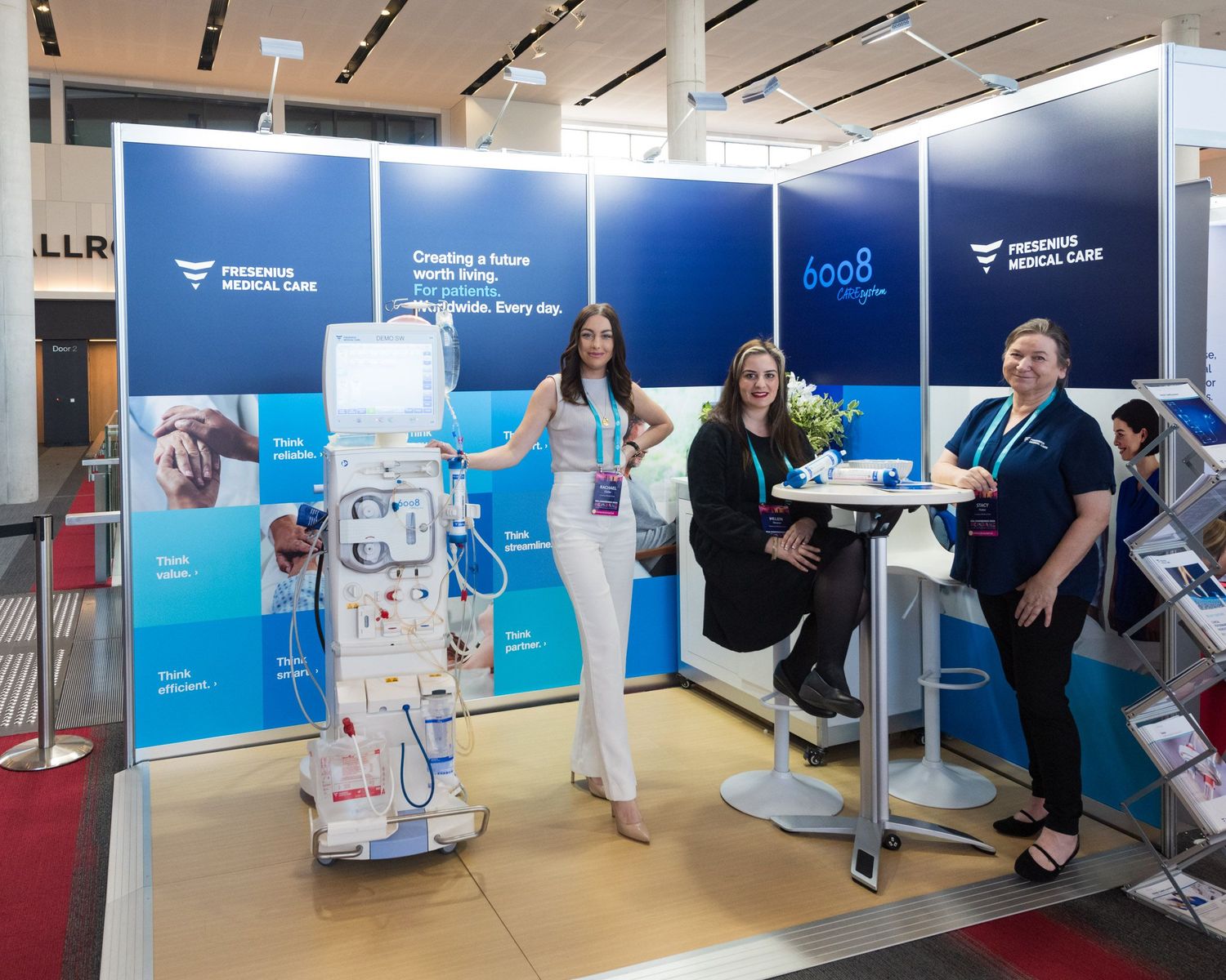Three women pose at Fresenius Medical Care exhibition booth with dialysis machine at medical conference.