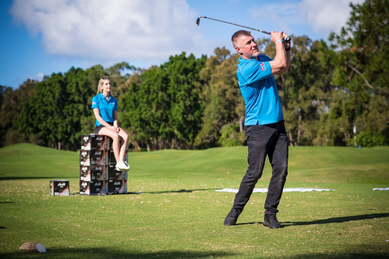 A man in a blue polo shirt swings a golf club on a sunny golf course while a child watches nearby.