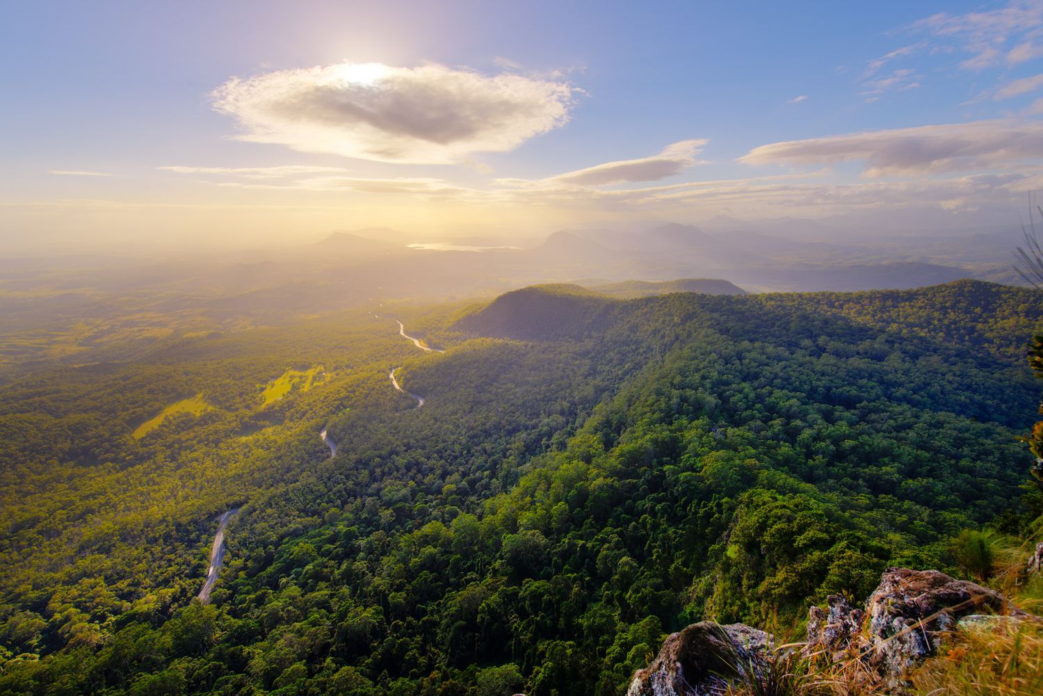 Aerial sunset view over a lush green mountain range with a winding road below.