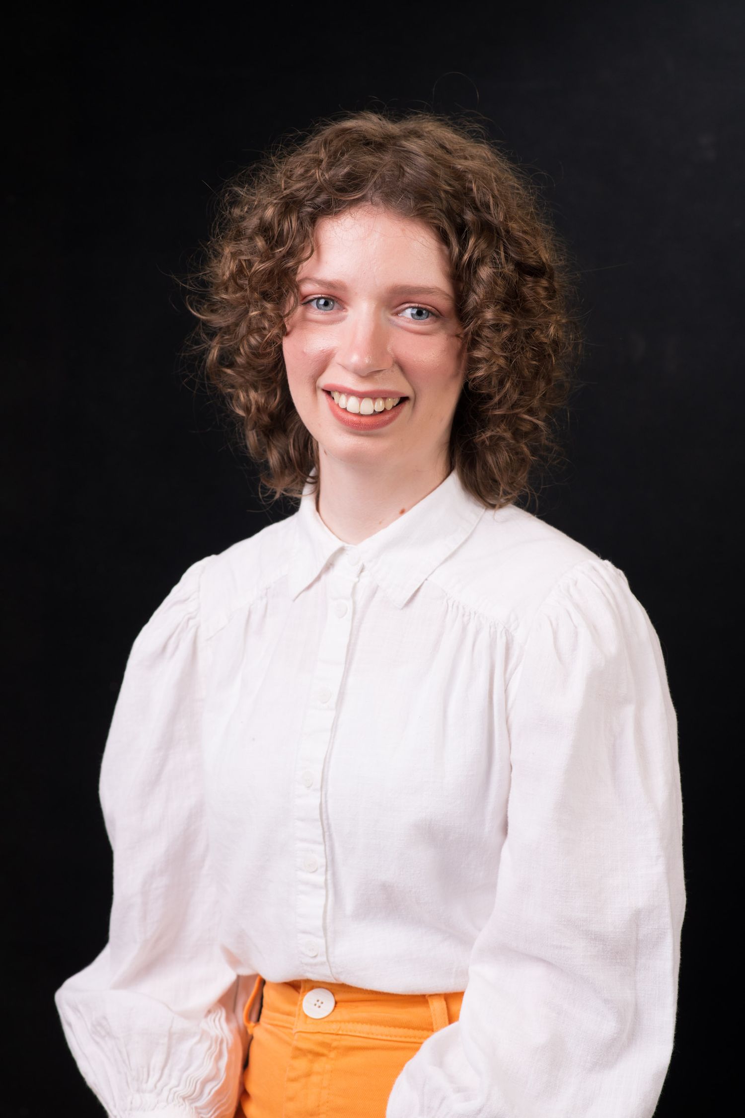 Young woman with curly hair smiling brightly wearing a white blouse and orange skirt.