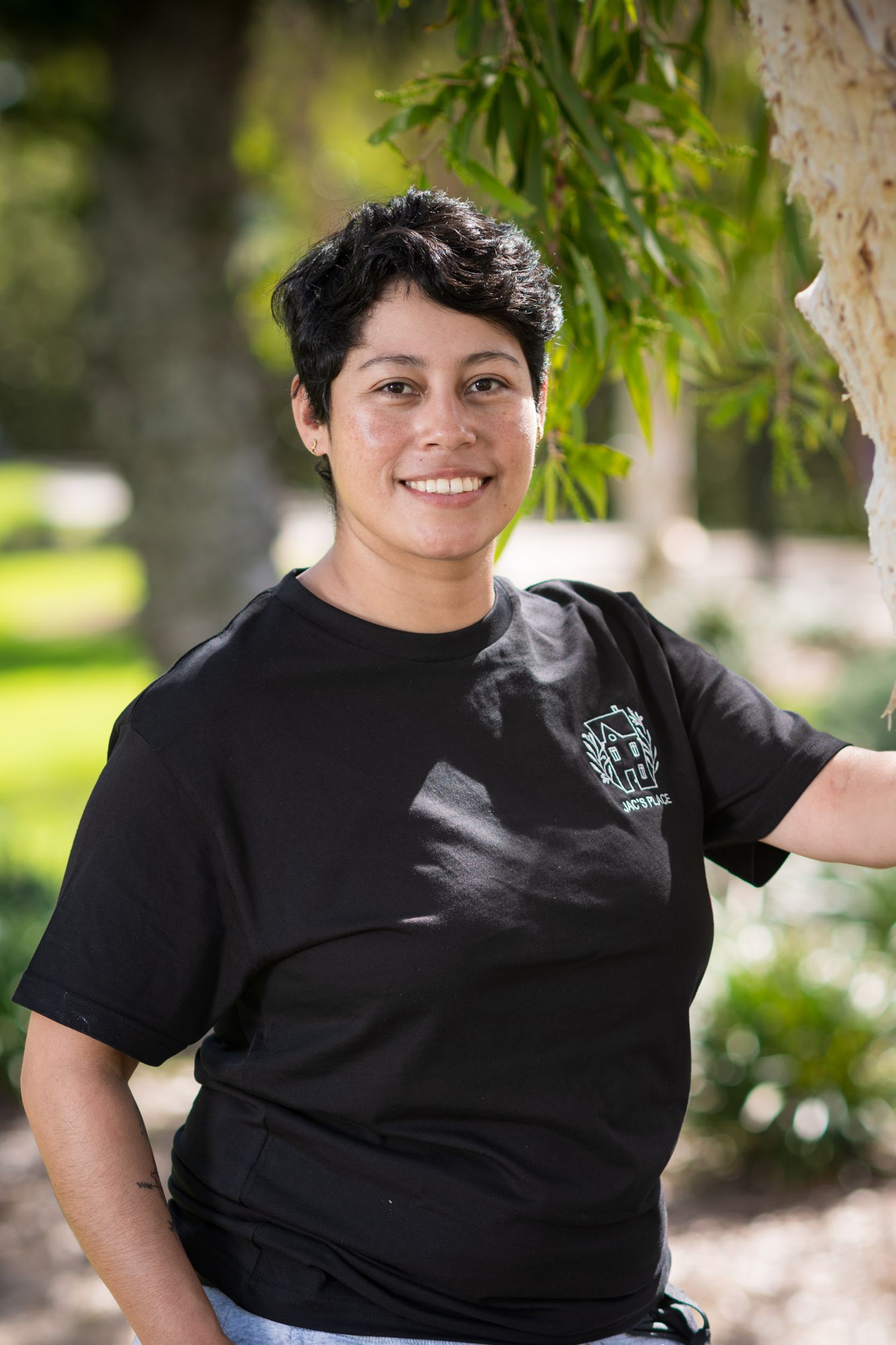 Smiling woman in black t-shirt leans against tree in sunny outdoor park setting.
