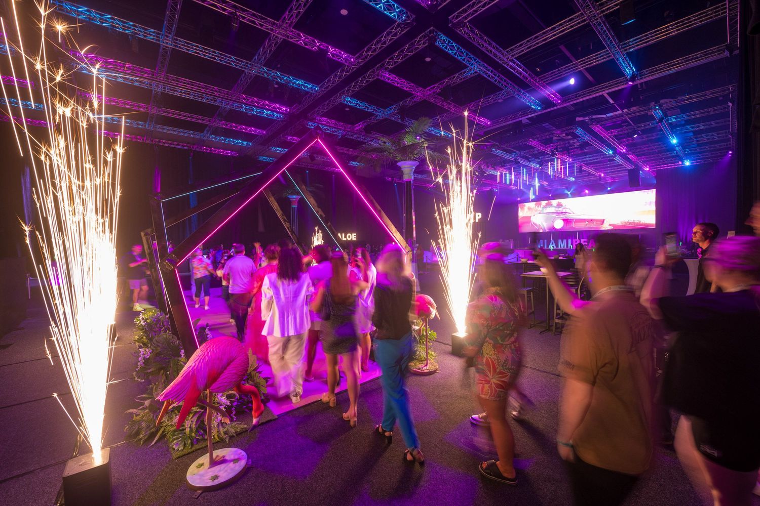 Guests entering a vibrant event through neon geometric arches with pyrotechnic sparklers and pink flamingo decor.