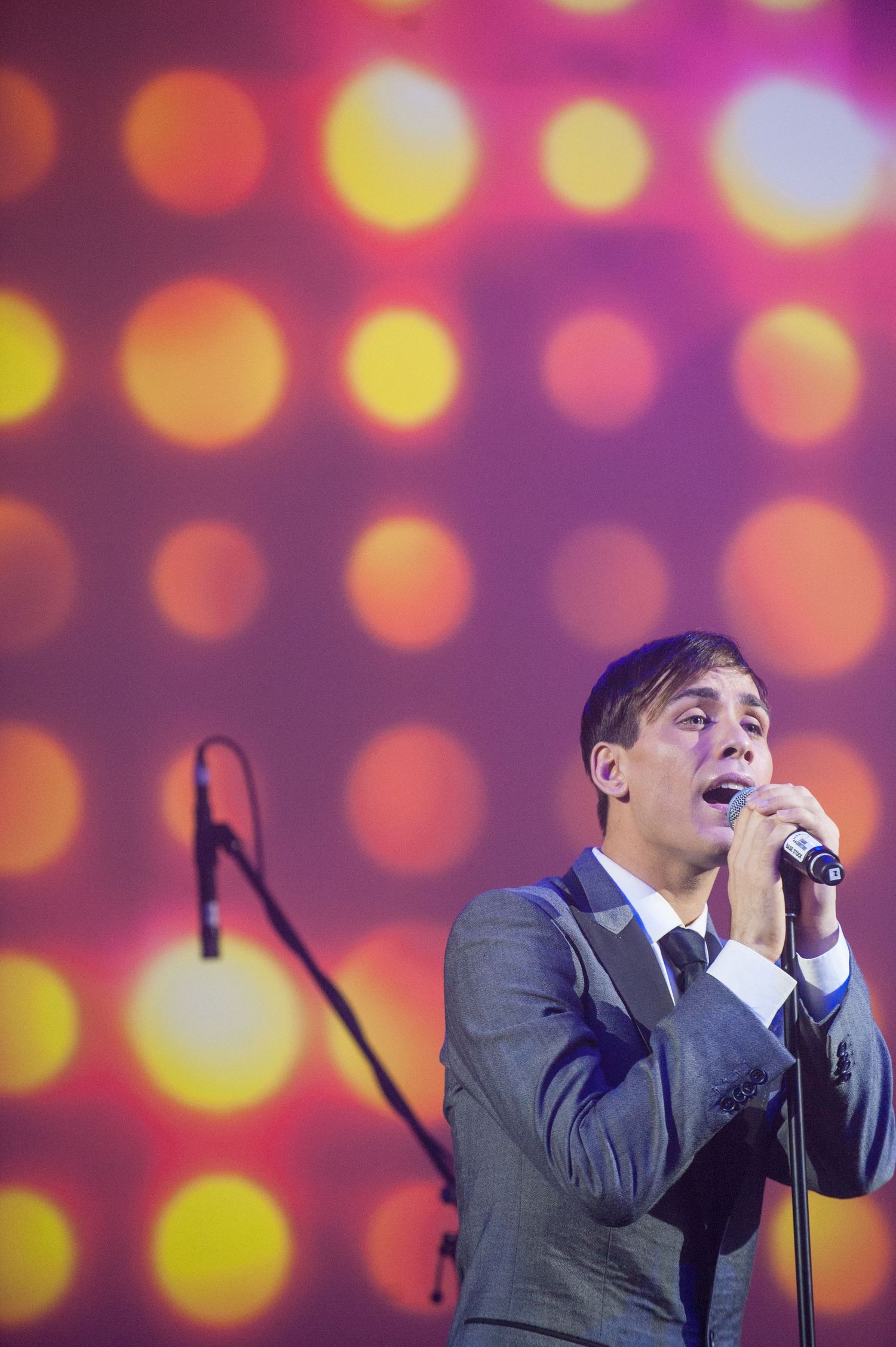 Male singer in grey suit performing on stage with colorful bokeh dot lighting backdrop.