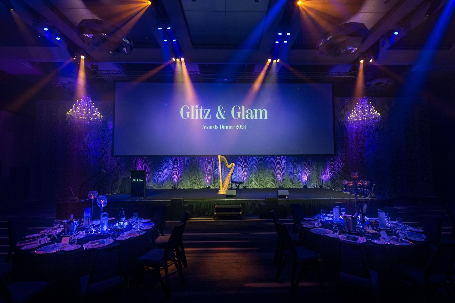 Elegant Glitz and Glam awards dinner venue with round tables, chandeliers, and dramatic stage lighting setup.