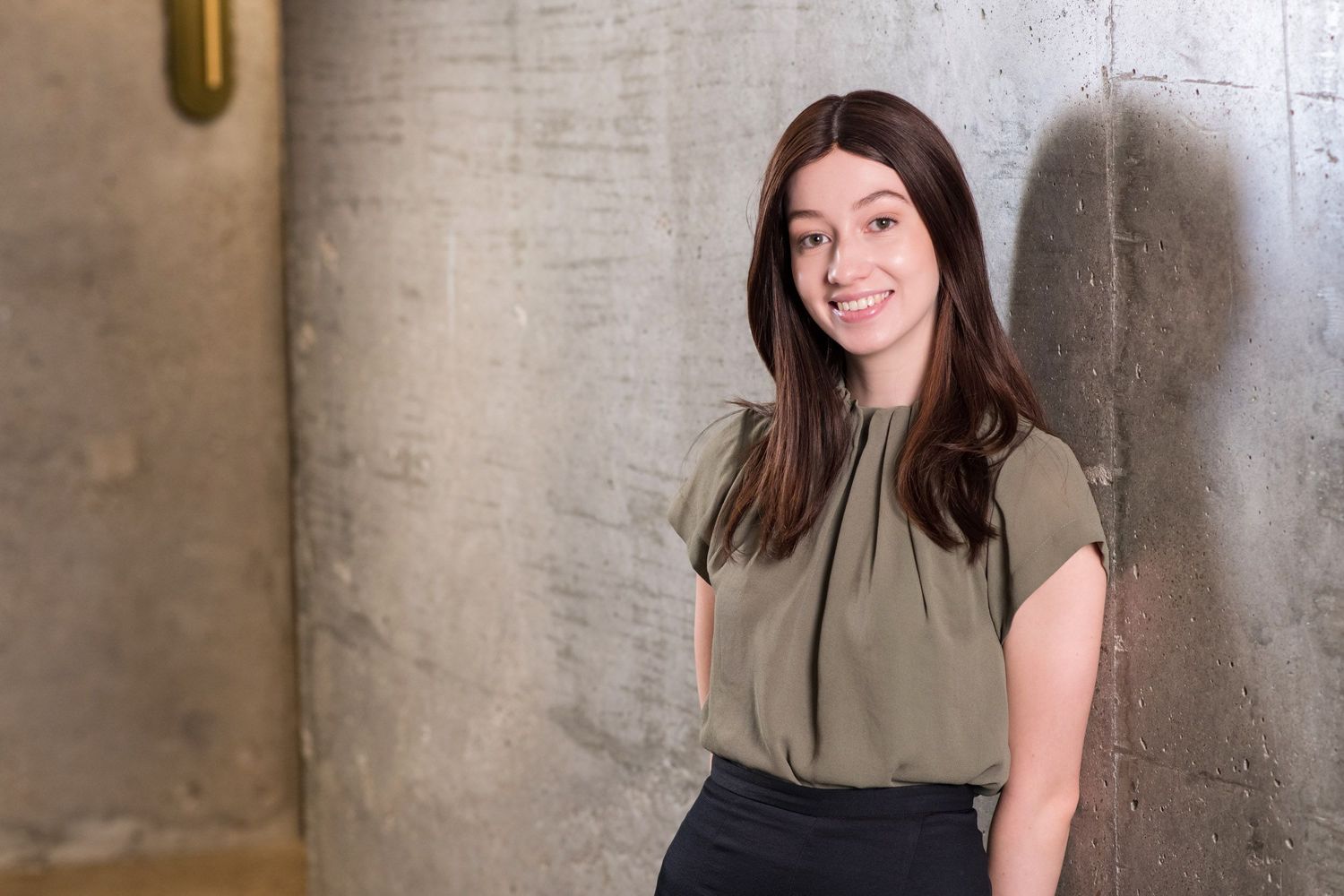 Young brunette woman in olive blouse smiling while leaning against a concrete wall.