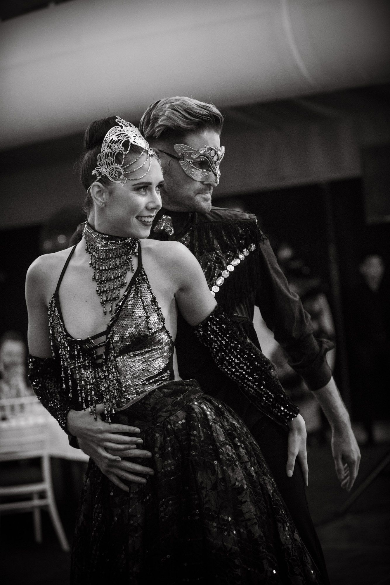 Black and white photo of elegantly dressed couple in masquerade masks dancing at a formal ball event.