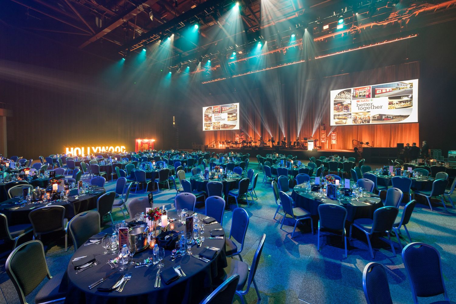 Elegant corporate gala dinner setup with blue-lit round tables, stage lighting, and large projection screens in a grand venue.