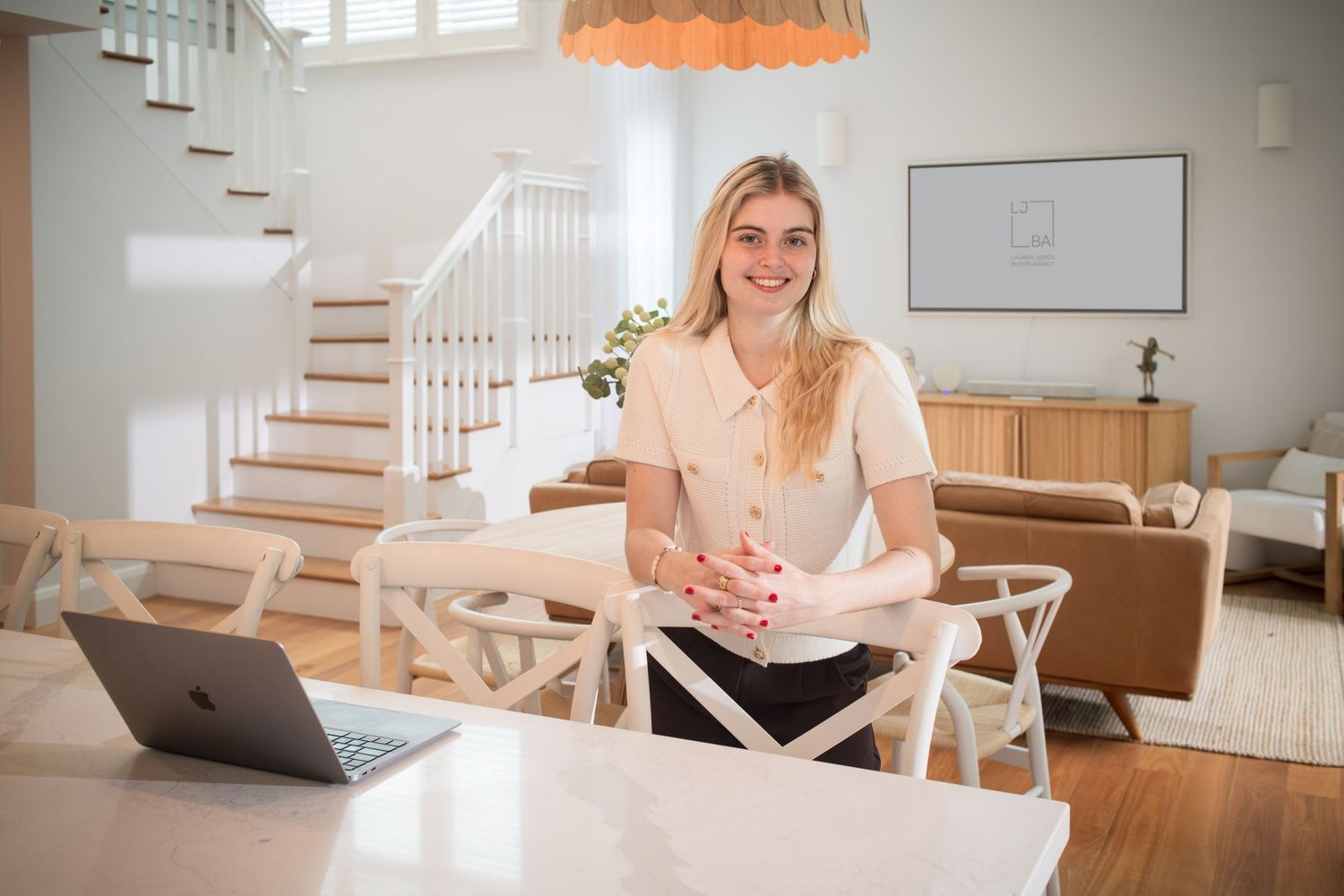 Smiling blonde woman leaning on a white dining table with a laptop in a modern home interior.