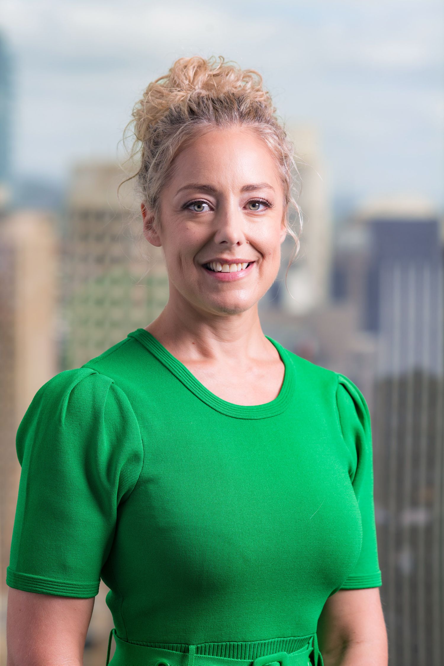 Smiling blonde woman in bright green top with city skyline visible in the blurred background.