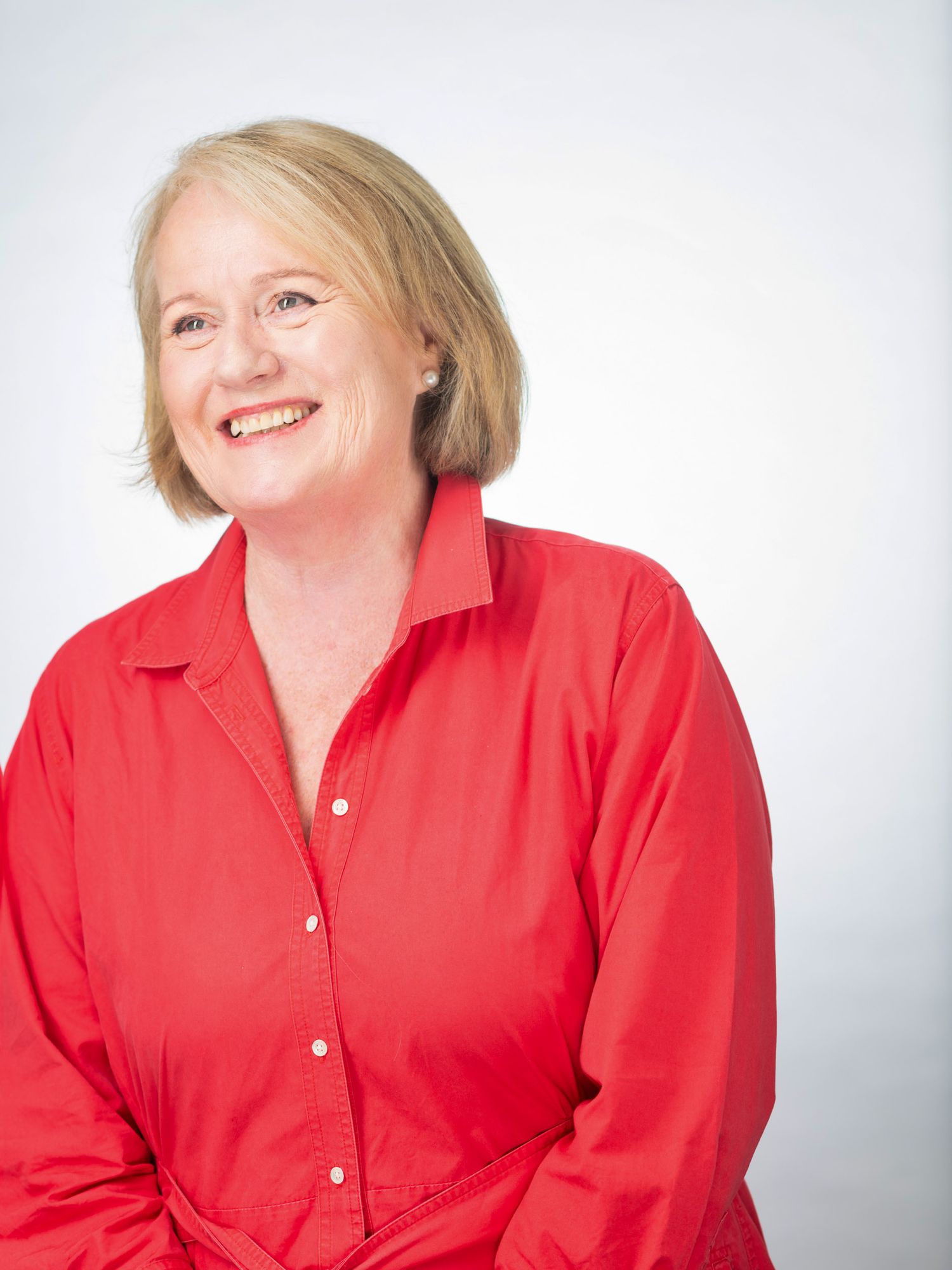 Smiling middle-aged woman with blonde hair wearing a bright red button-up shirt against a white background.