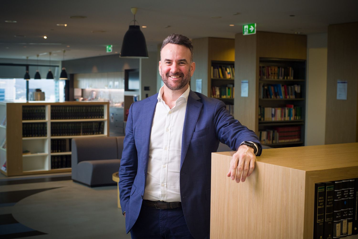 Confident man in blue blazer leans on wooden counter smiling in modern library setting.