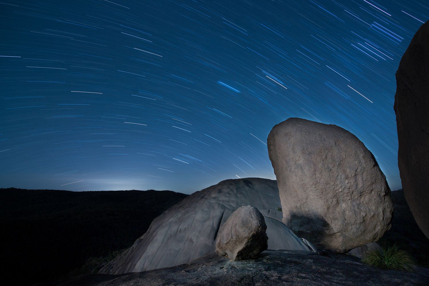 Long exposure star trails arc over illuminated desert boulders in dark night sky landscape.