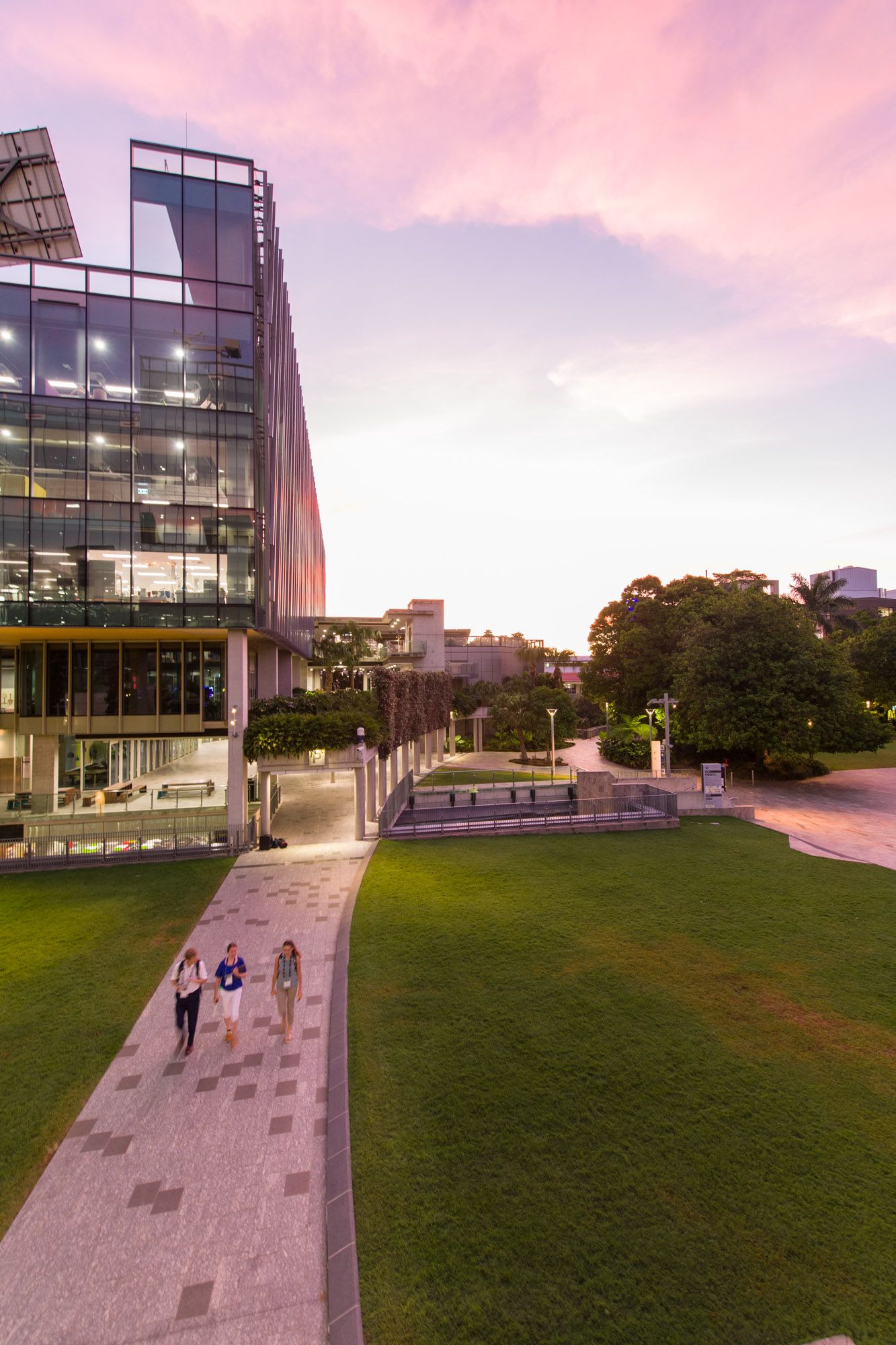 Students walk past a contemporary glass campus building at dusk under a colorful pink sunset sky.