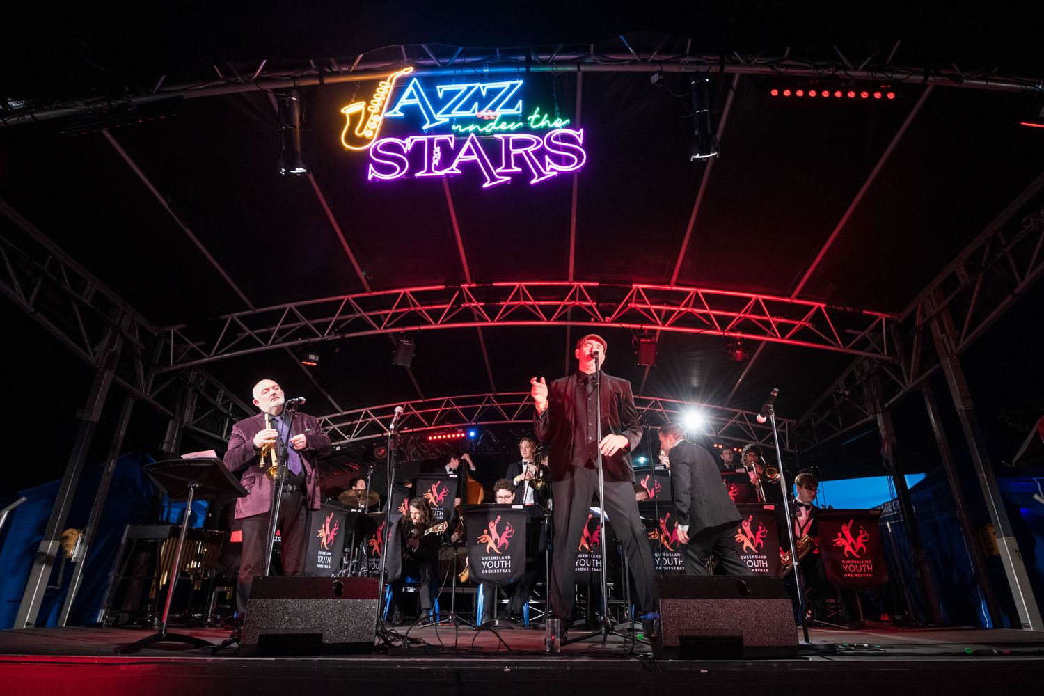 Jazz band performing live on stage under Jazz Under the Stars neon sign at night event.