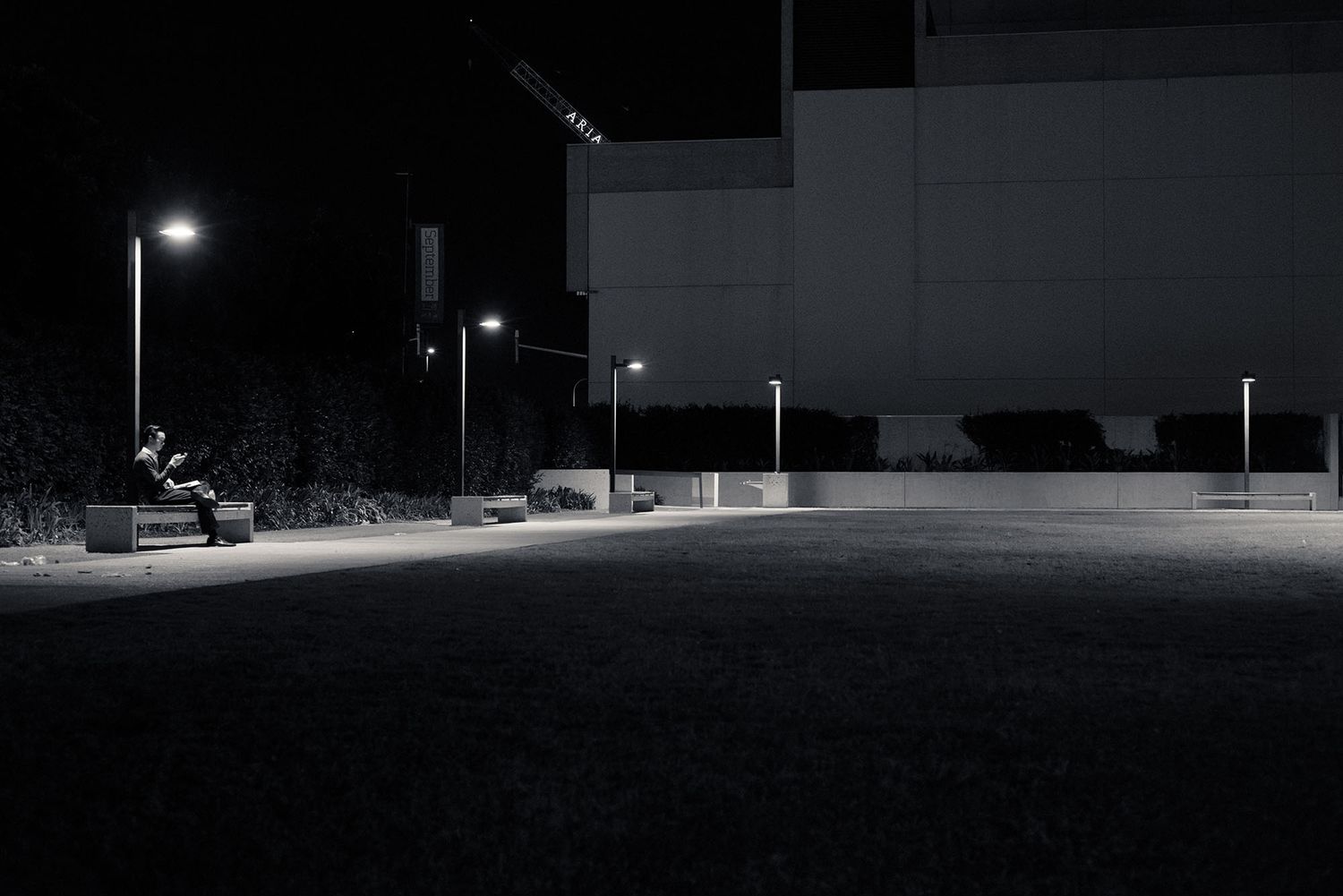Empty outdoor plaza lit by street lamps at night beside a large building in monochrome.