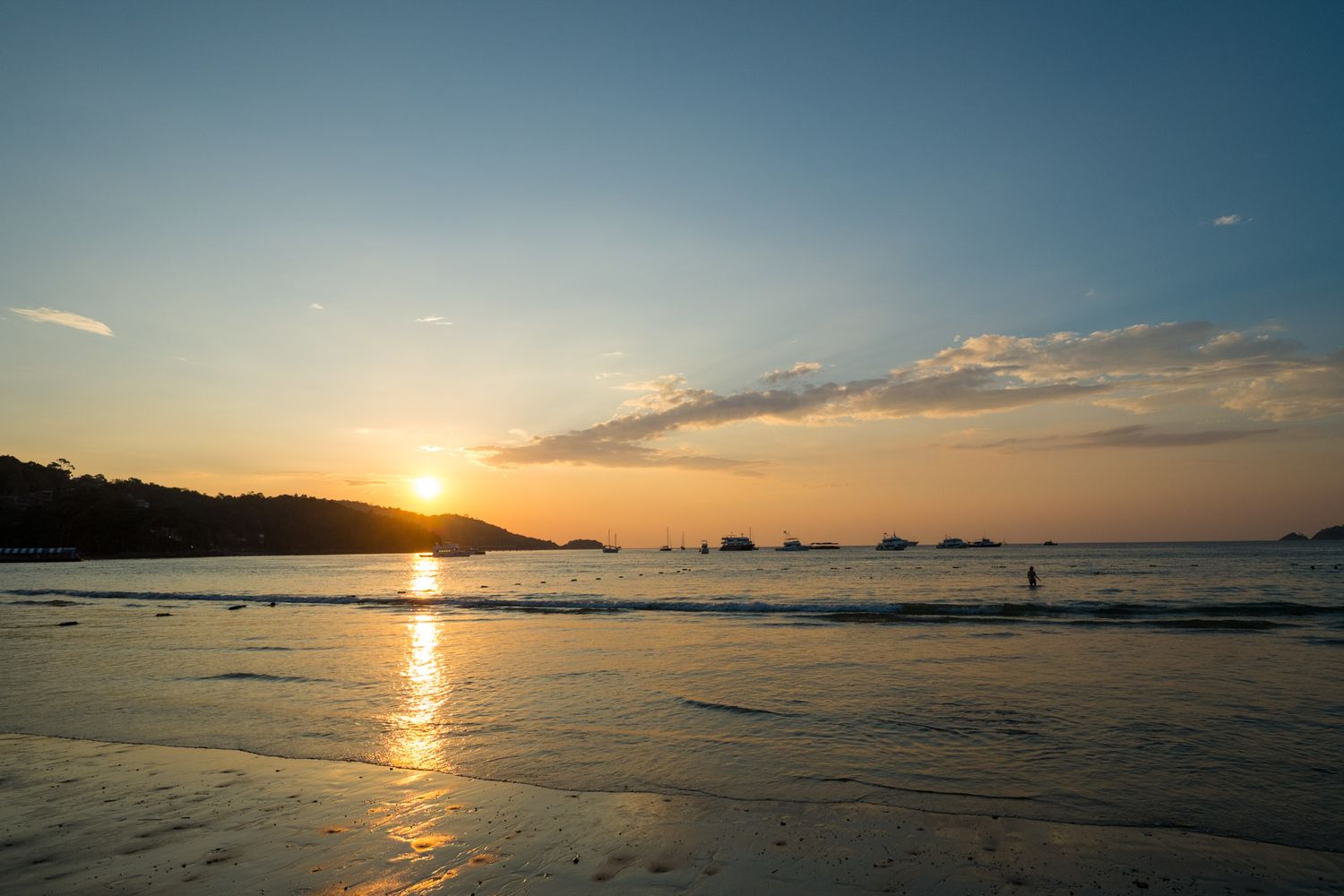 Golden sunset reflects on wet sand as a lone figure stands at the shoreline of a tropical beach.