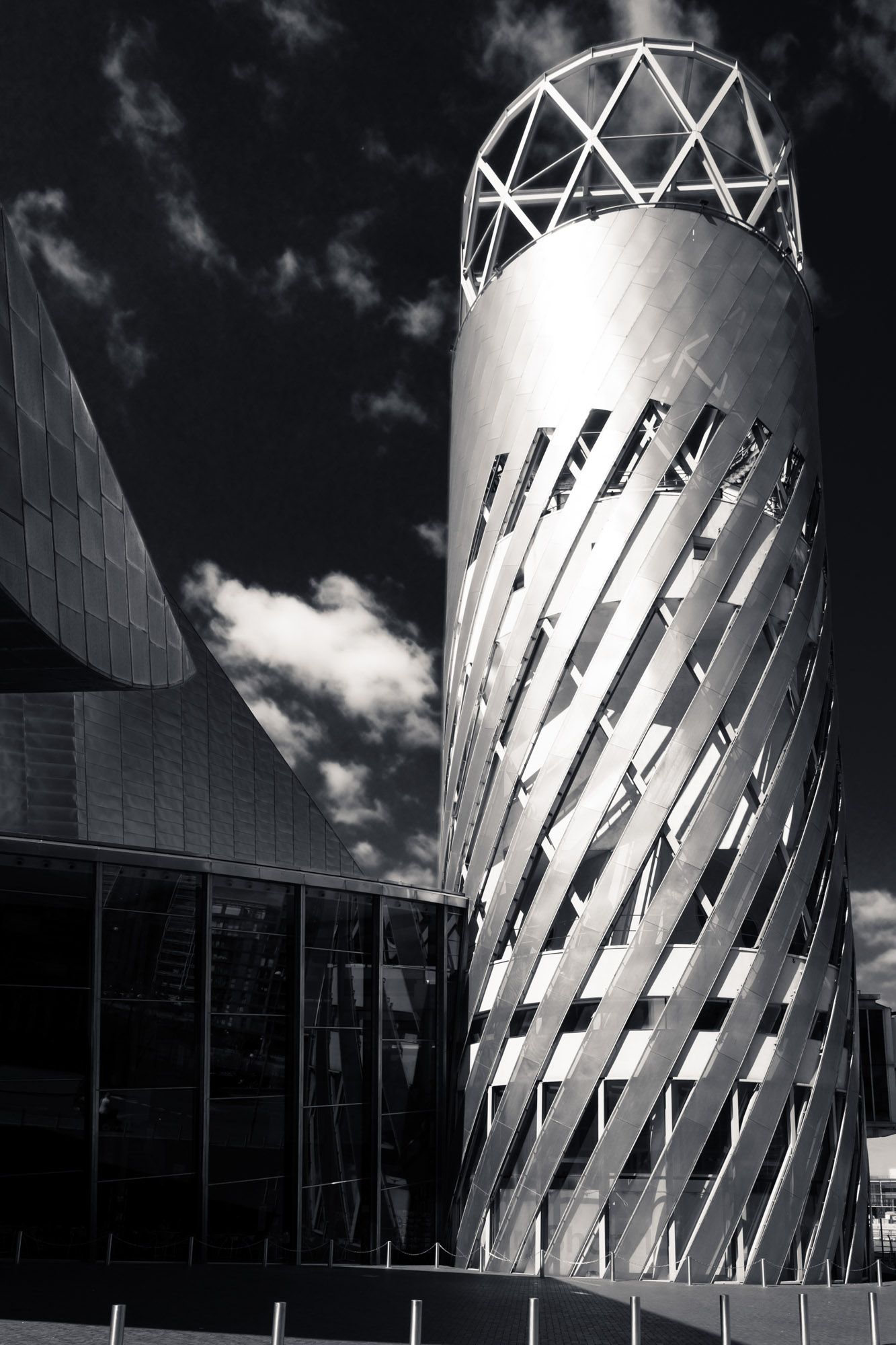 Black and white photo of a modern metallic cylindrical tower with diagonal lattice patterns against cloudy sky.