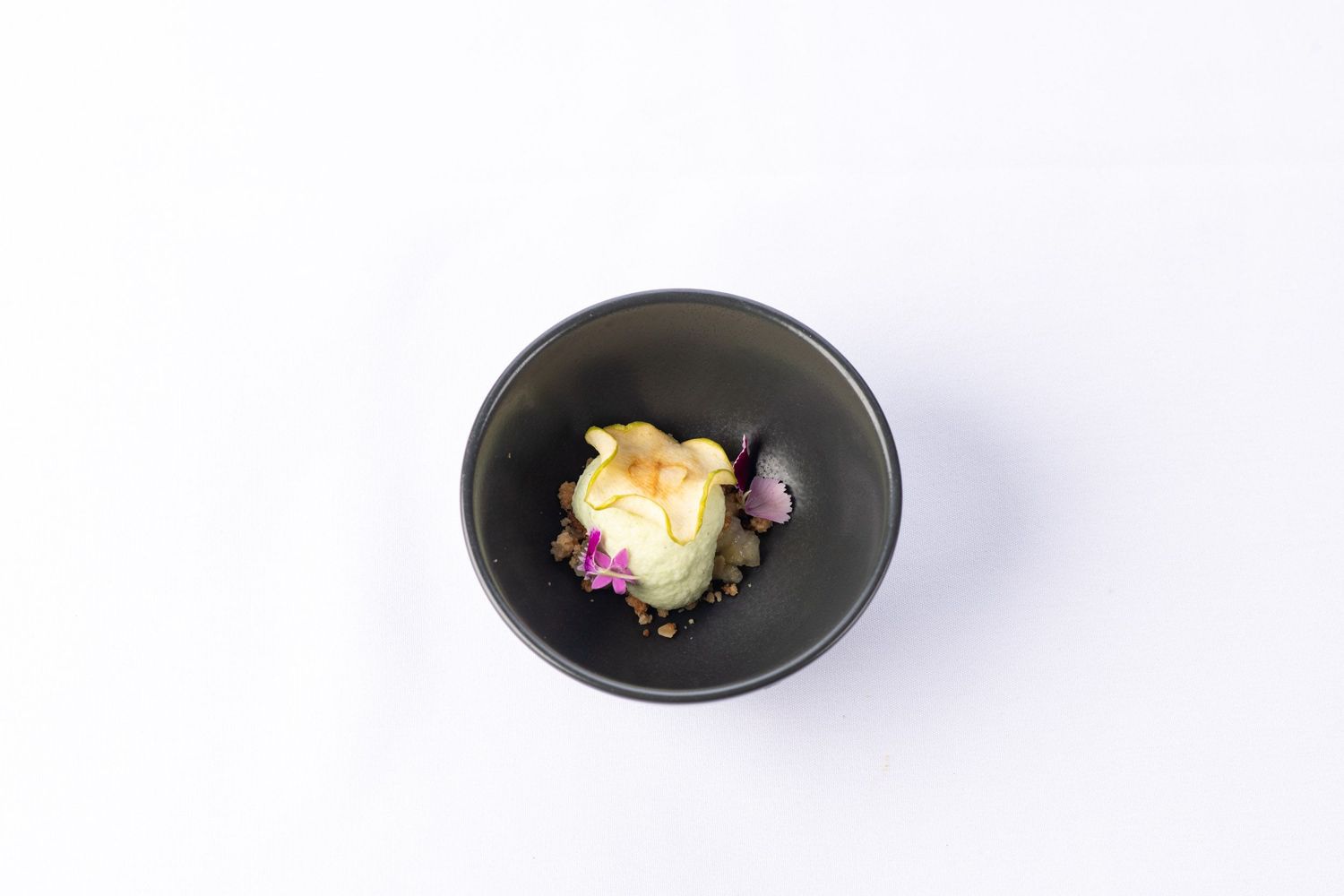 Fine dining dessert with golden pastry and purple flower garnish served in a black ceramic bowl.