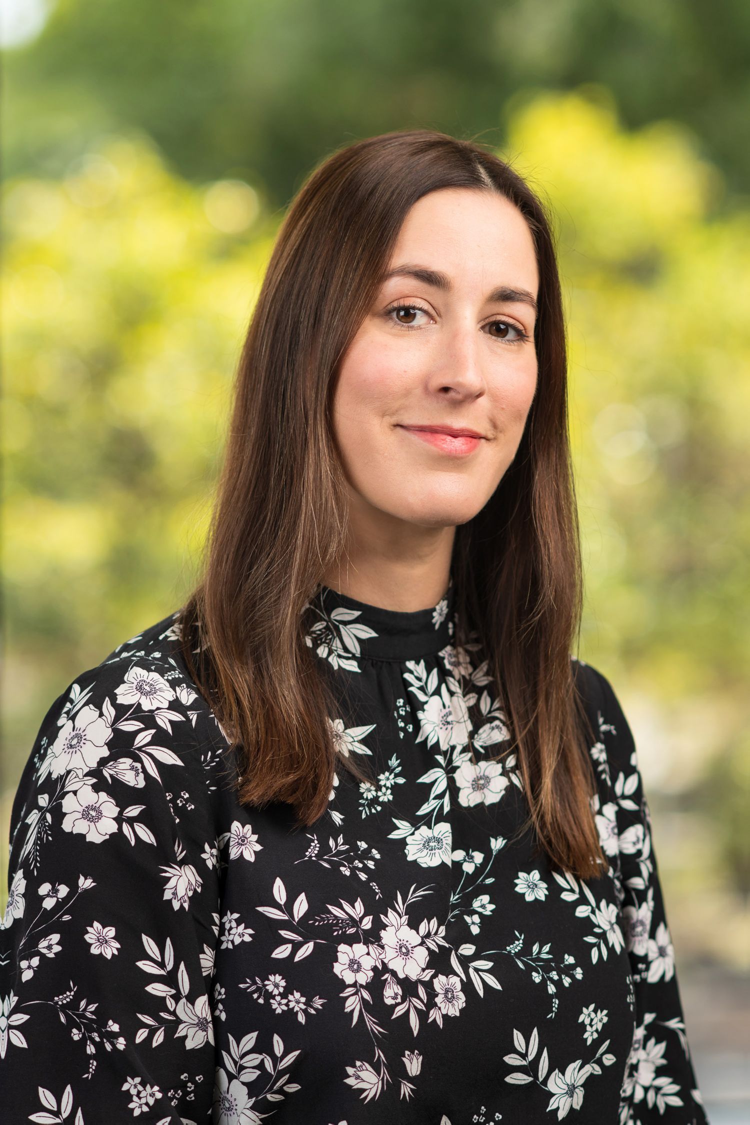 Woman with brown hair smiling outdoors wearing a black floral dress, professional headshot with green bokeh background.