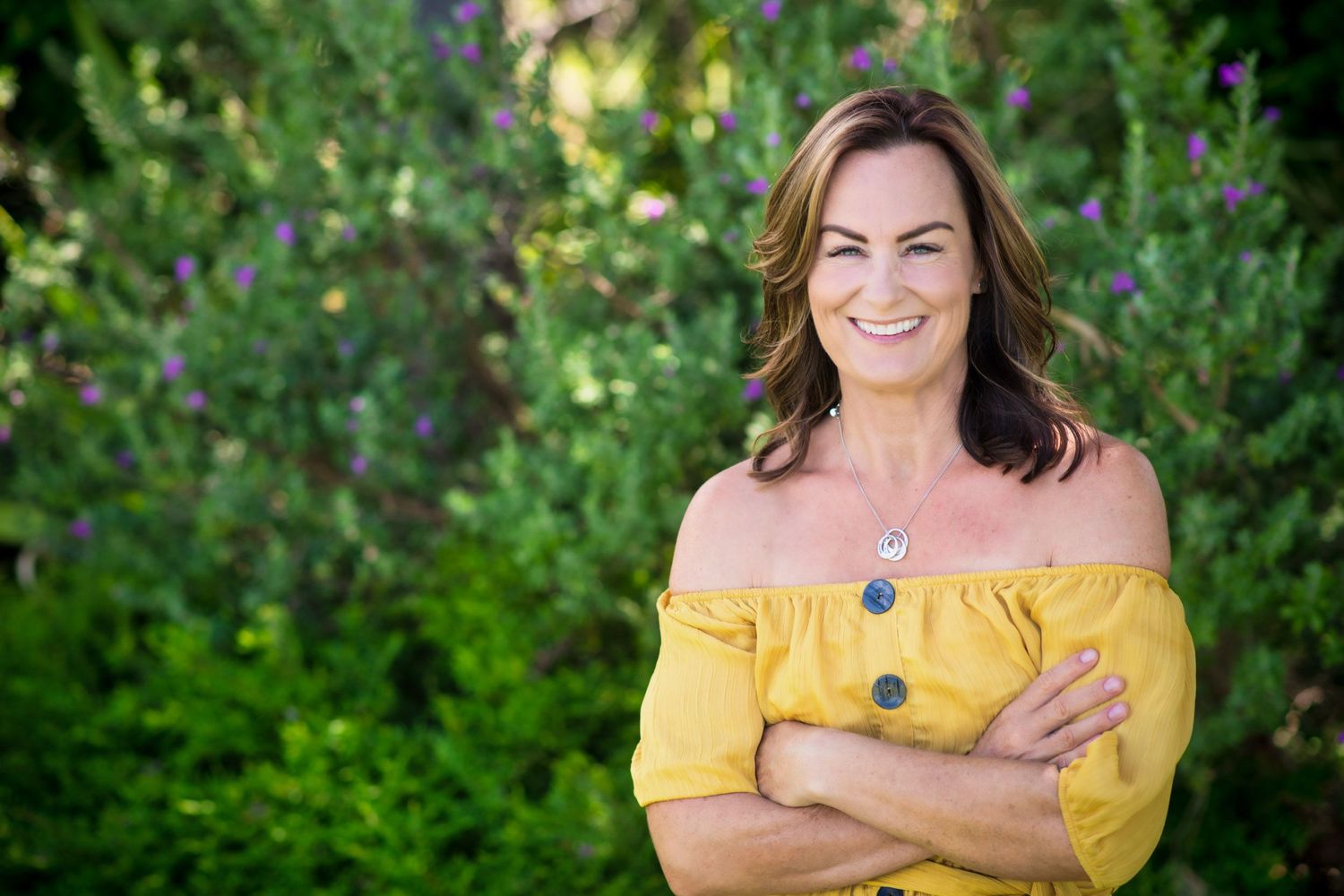 Smiling woman in yellow off-shoulder top stands with arms crossed in lush garden setting.