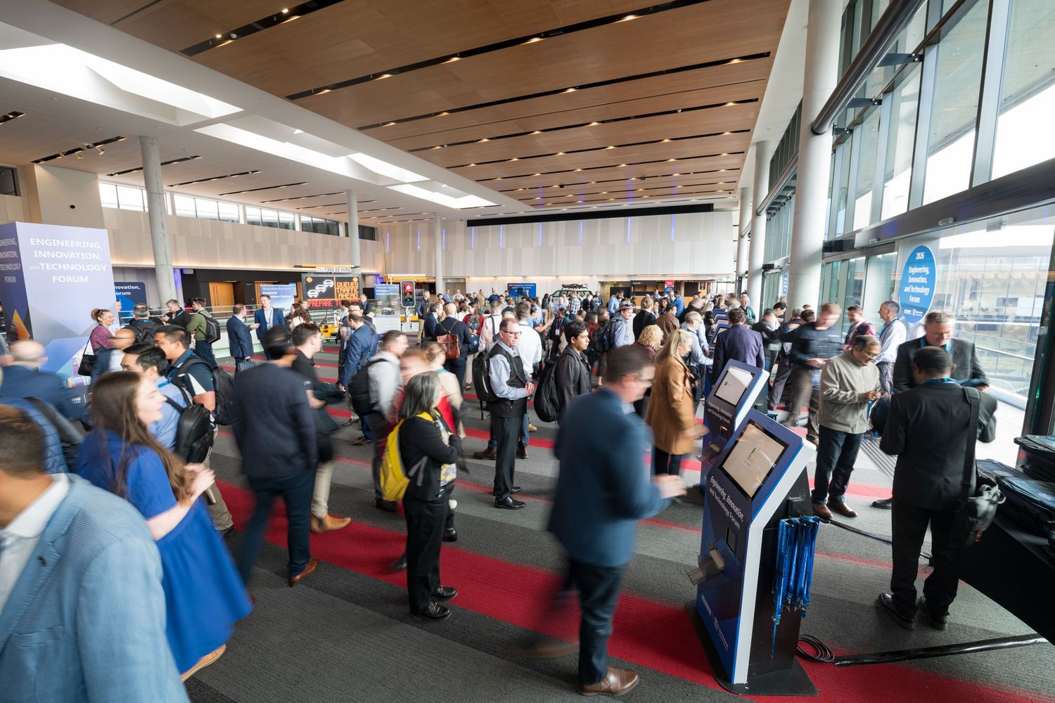 Busy conference lobby with attendees networking near registration kiosks and information displays.