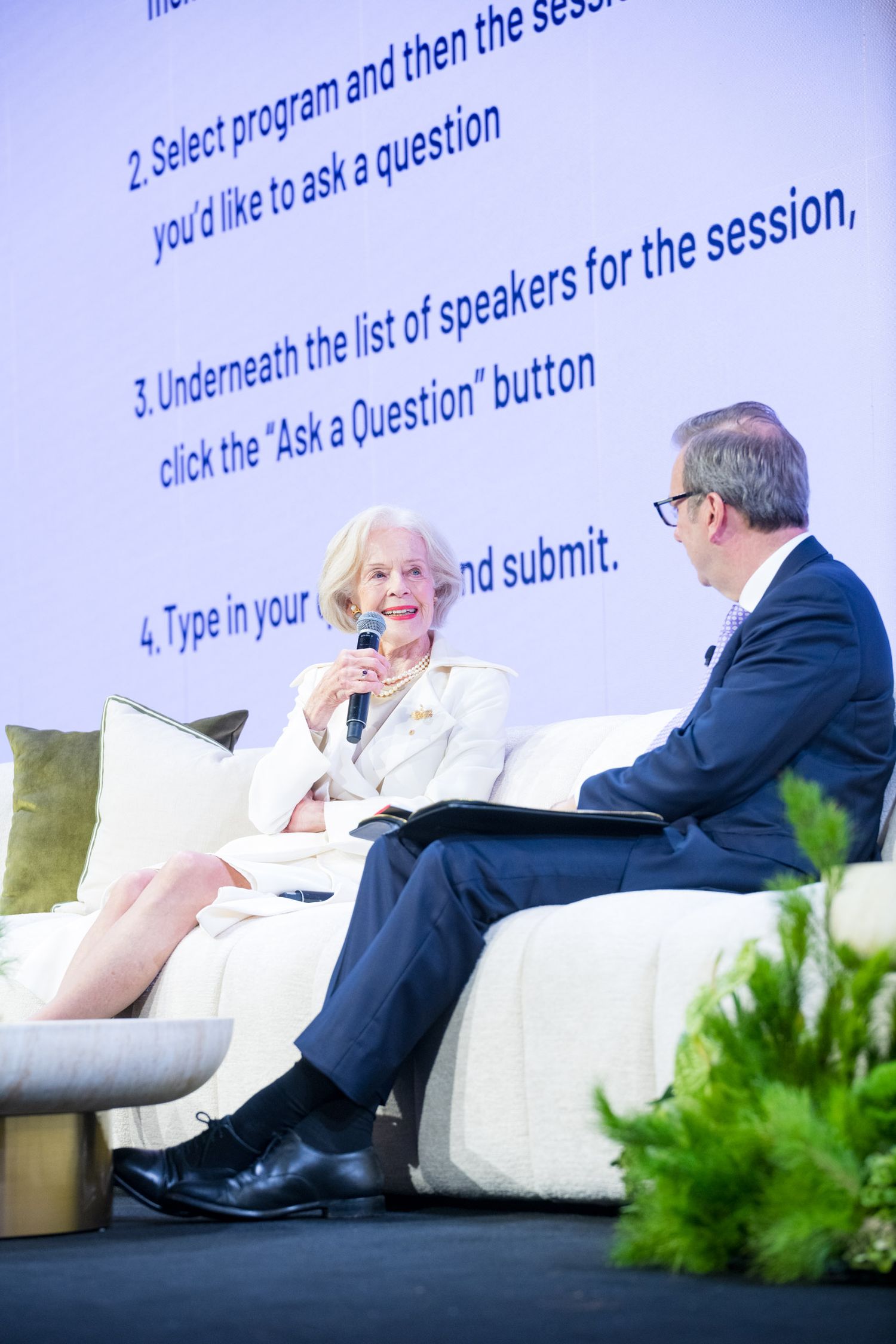 Two speakers conversing on stage at conference with Q&A instructions displayed on large screen.