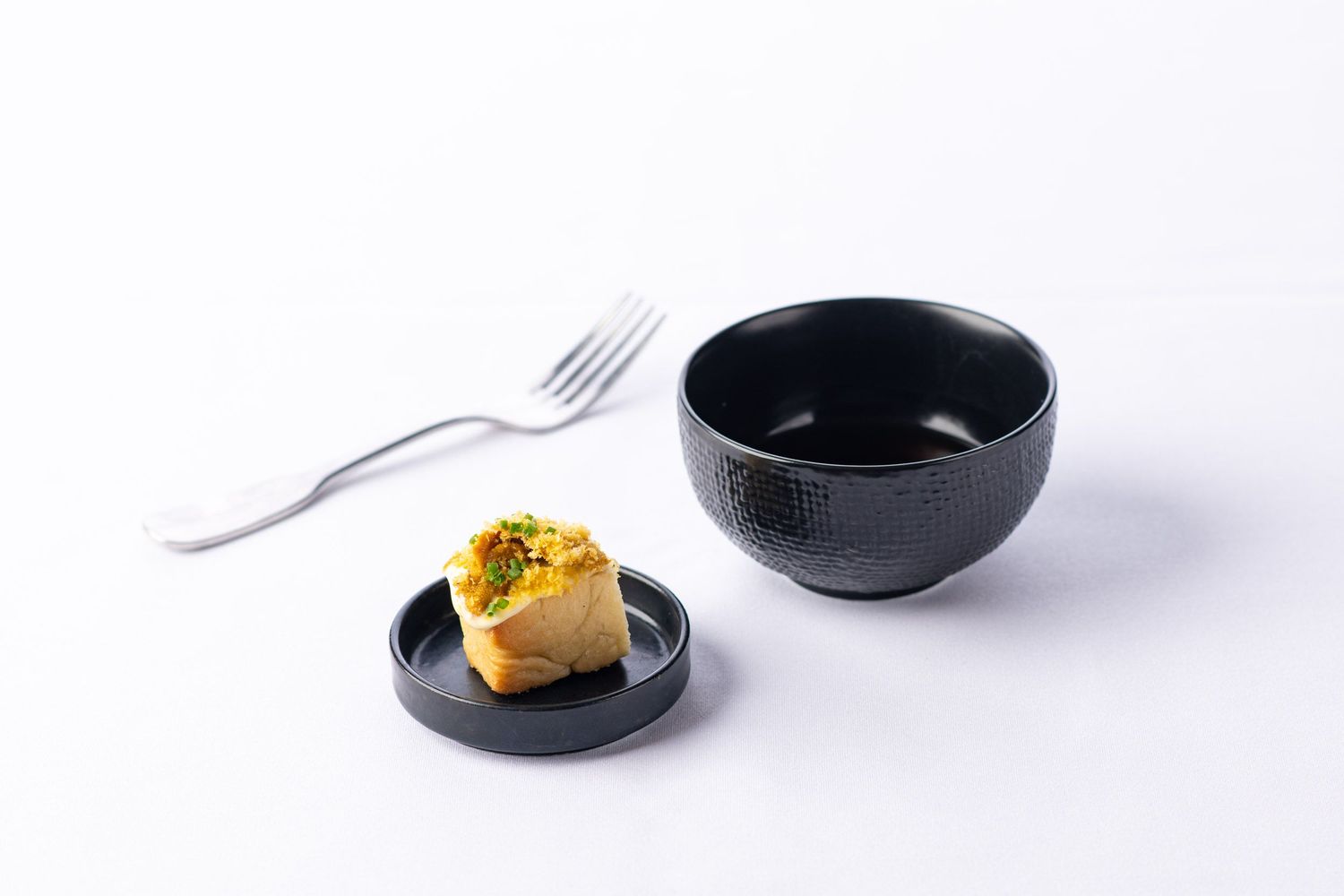 Black ceramic bowl with small plate holding a golden pastry bite and silver fork on white background.