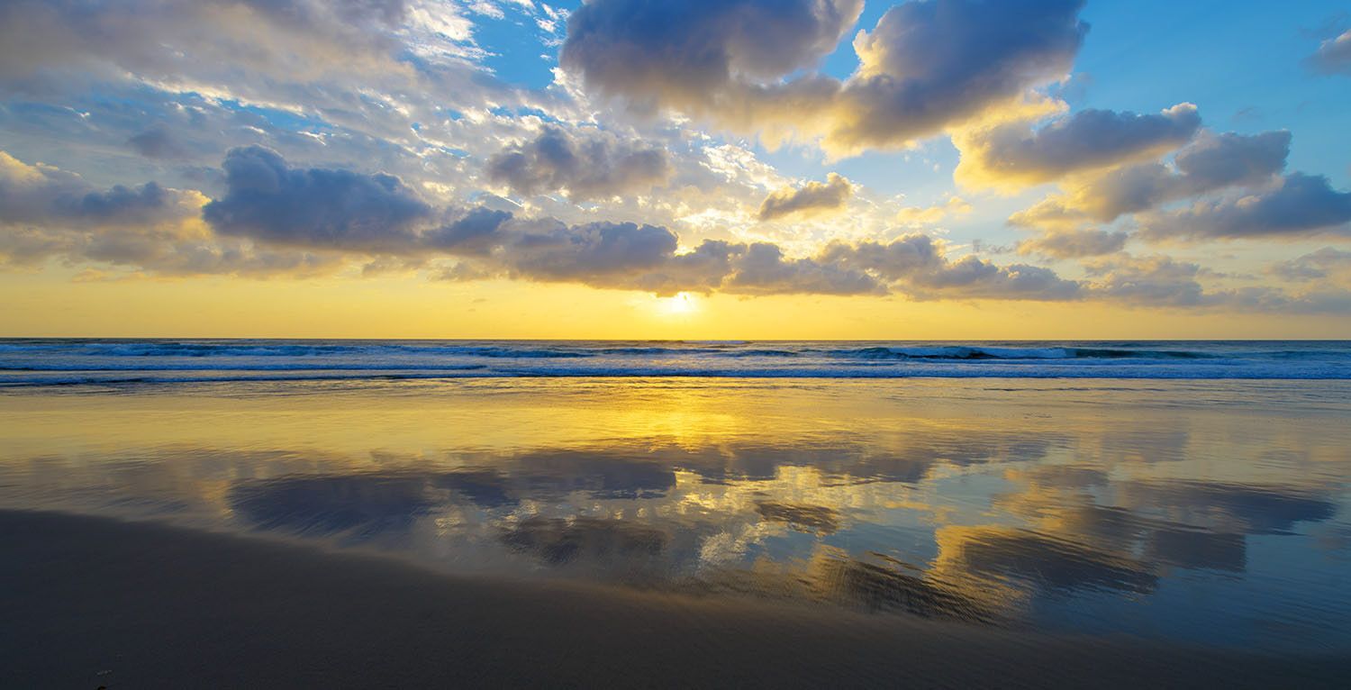 Breathtaking beach sunset reflected on wet sand with dramatic golden clouds over ocean waves.