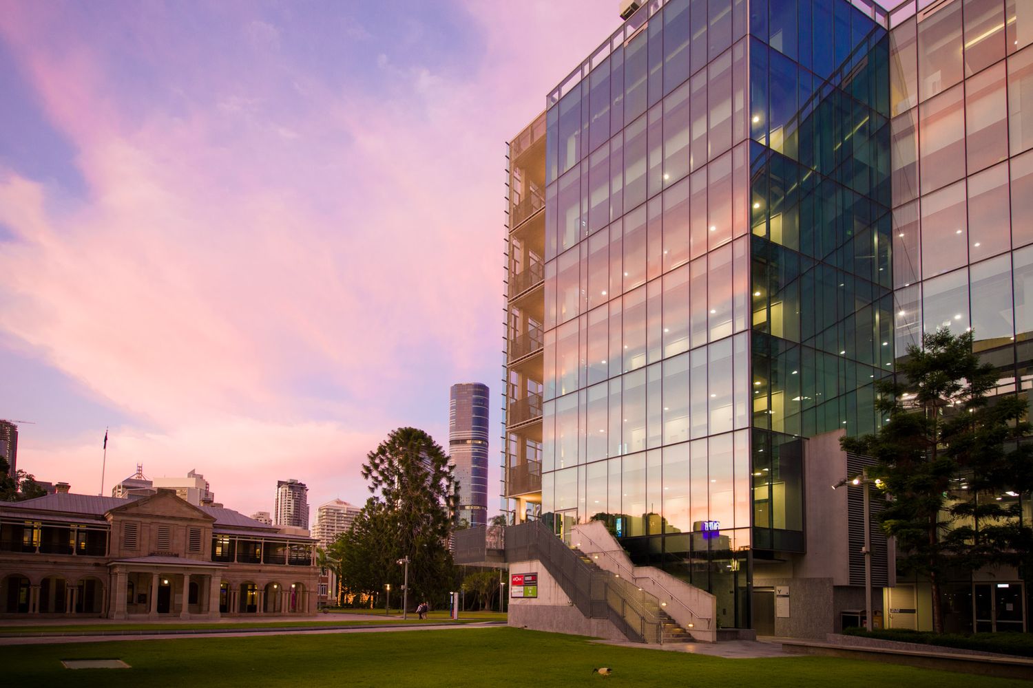Modern glass university building reflects a stunning pink sunset sky beside historic Brisbane architecture.