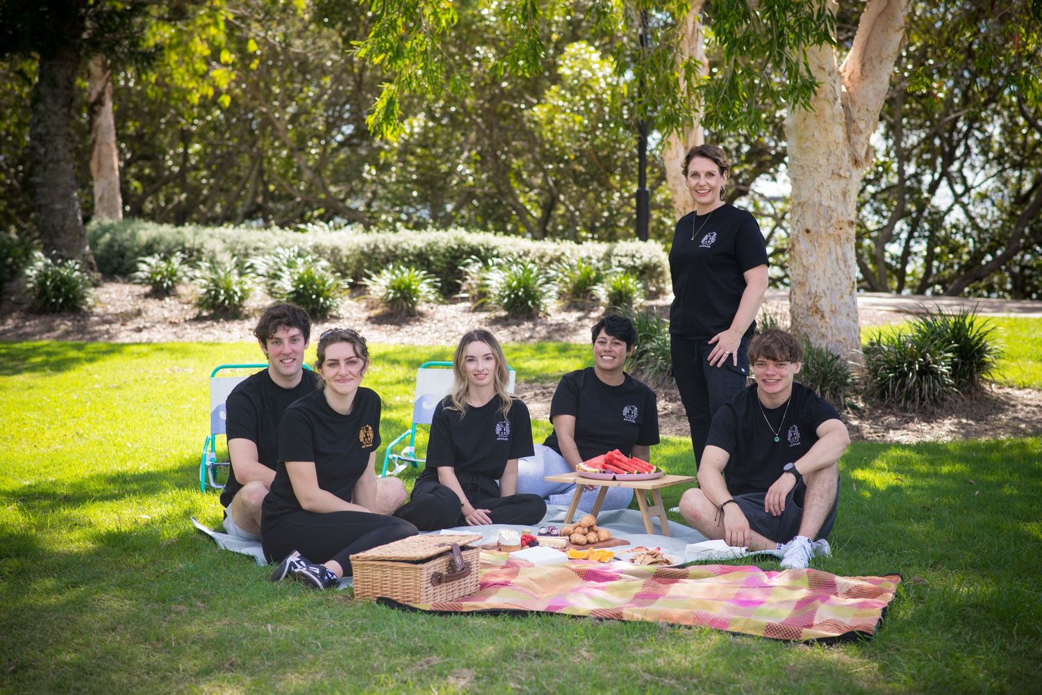 Group of six young people in black shirts enjoying a picnic on green grass in a scenic park.