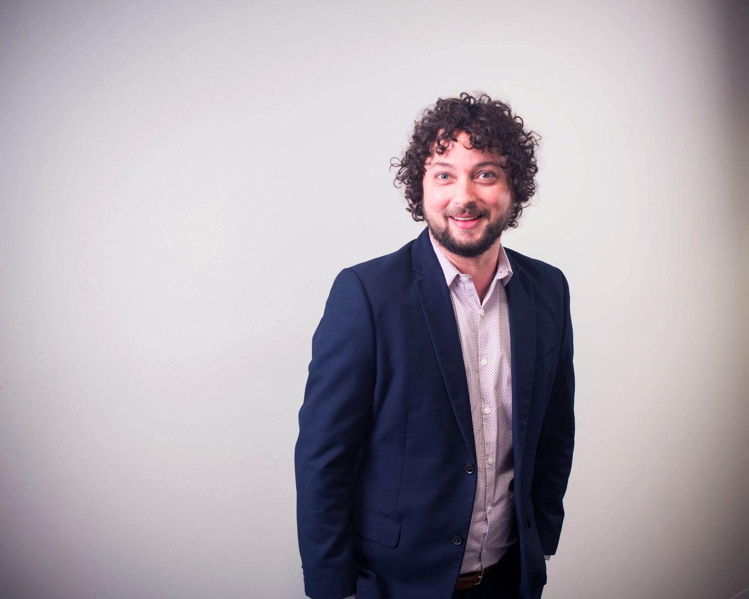 Smiling man with curly hair wearing navy blazer and open-collar shirt poses against grey background.