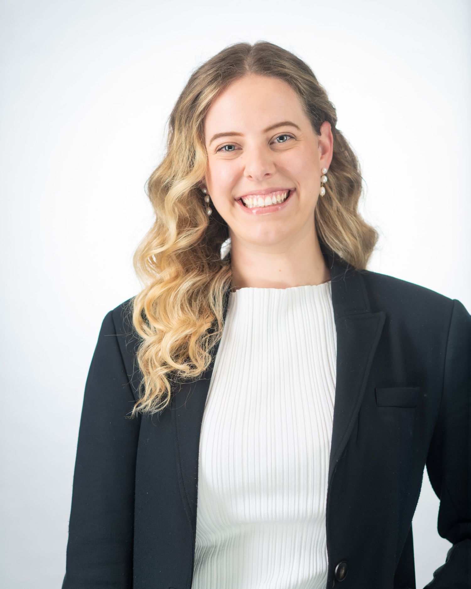 Smiling woman with curly blonde hair in black blazer, professional business portrait on light background.