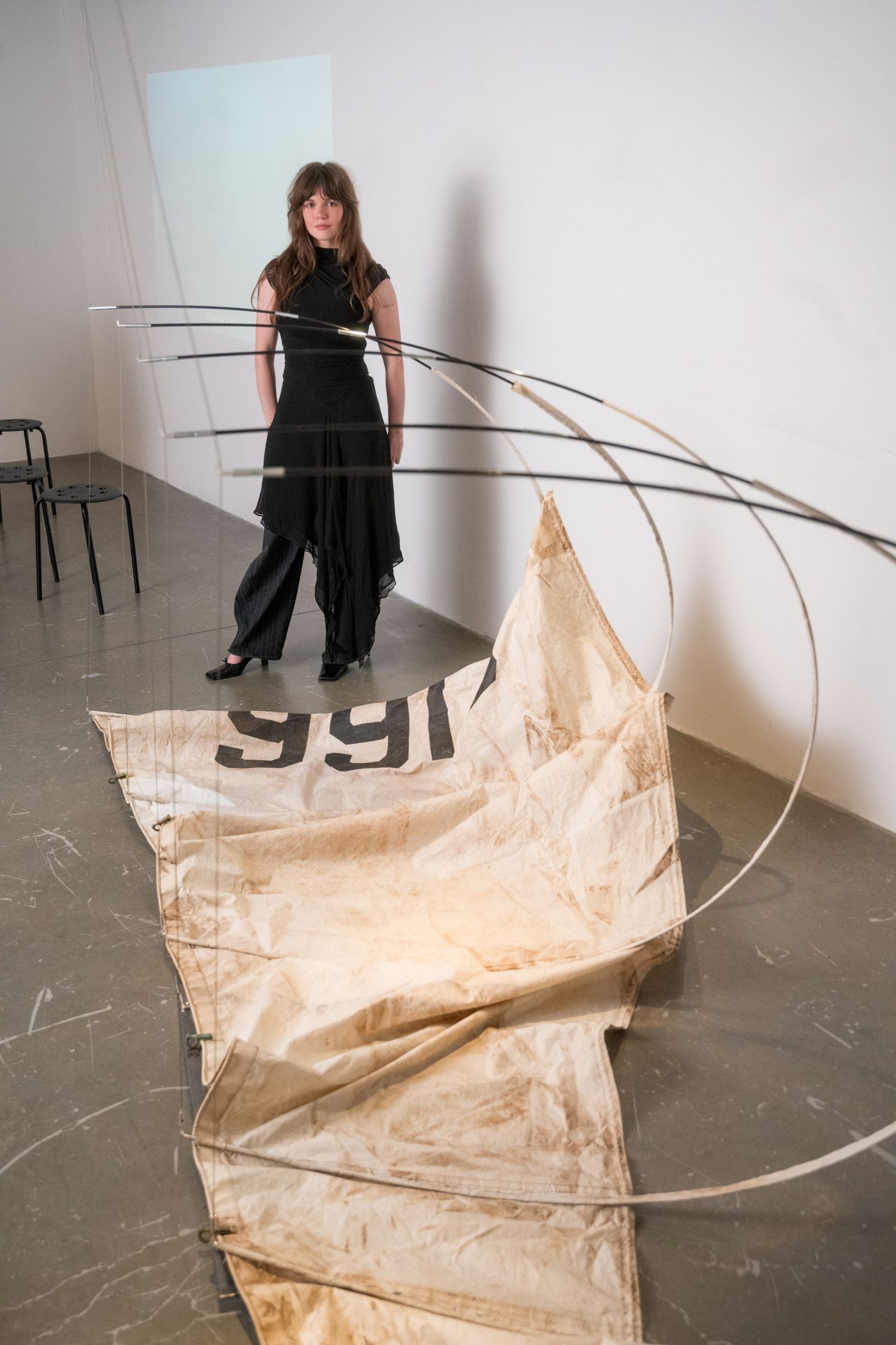 Woman in black outfit standing behind curved wire sculpture with weathered canvas in gallery.