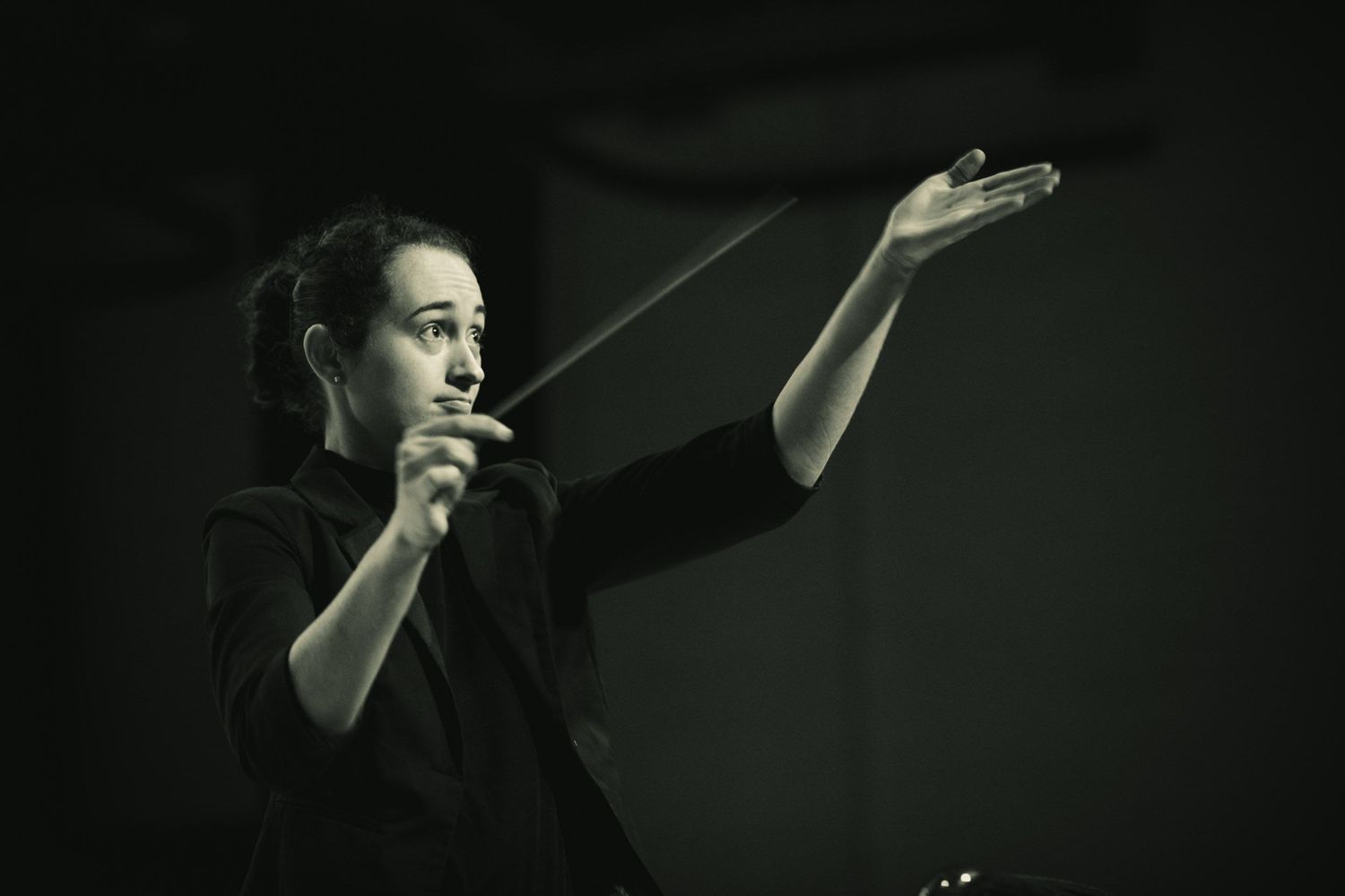 Female conductor holding a baton mid-performance in a dramatic black and white photo.