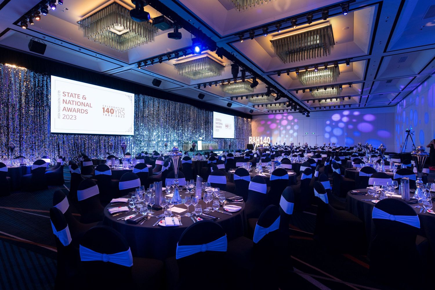 Corporate awards gala venue with round tables, black chair covers, and illuminated screens at State National Awards 2023.