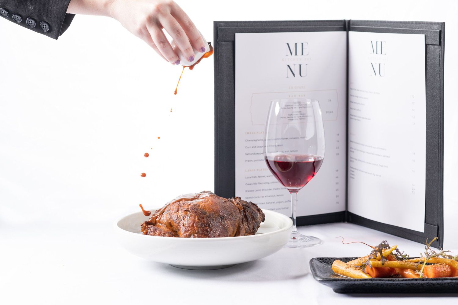 Gourmet roasted meat dish being sauced by hand with red wine glass and menu in background.