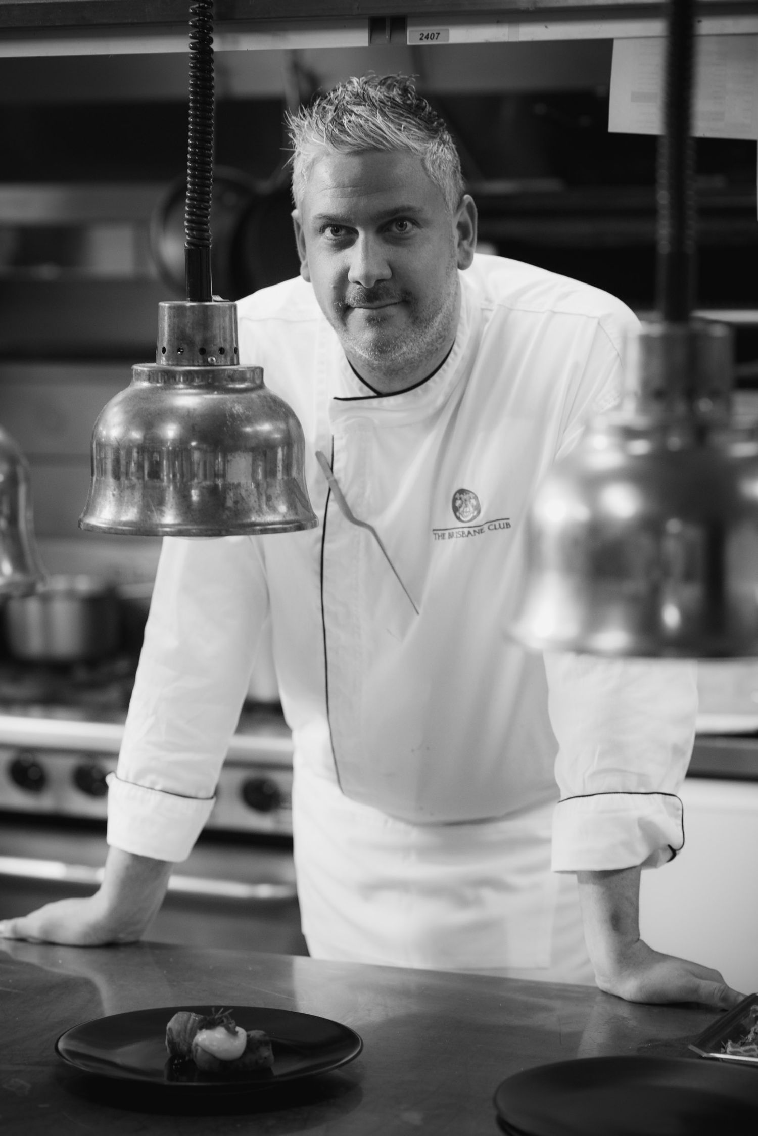 Black and white portrait of a chef in white uniform leaning over a kitchen counter with plated dishes.