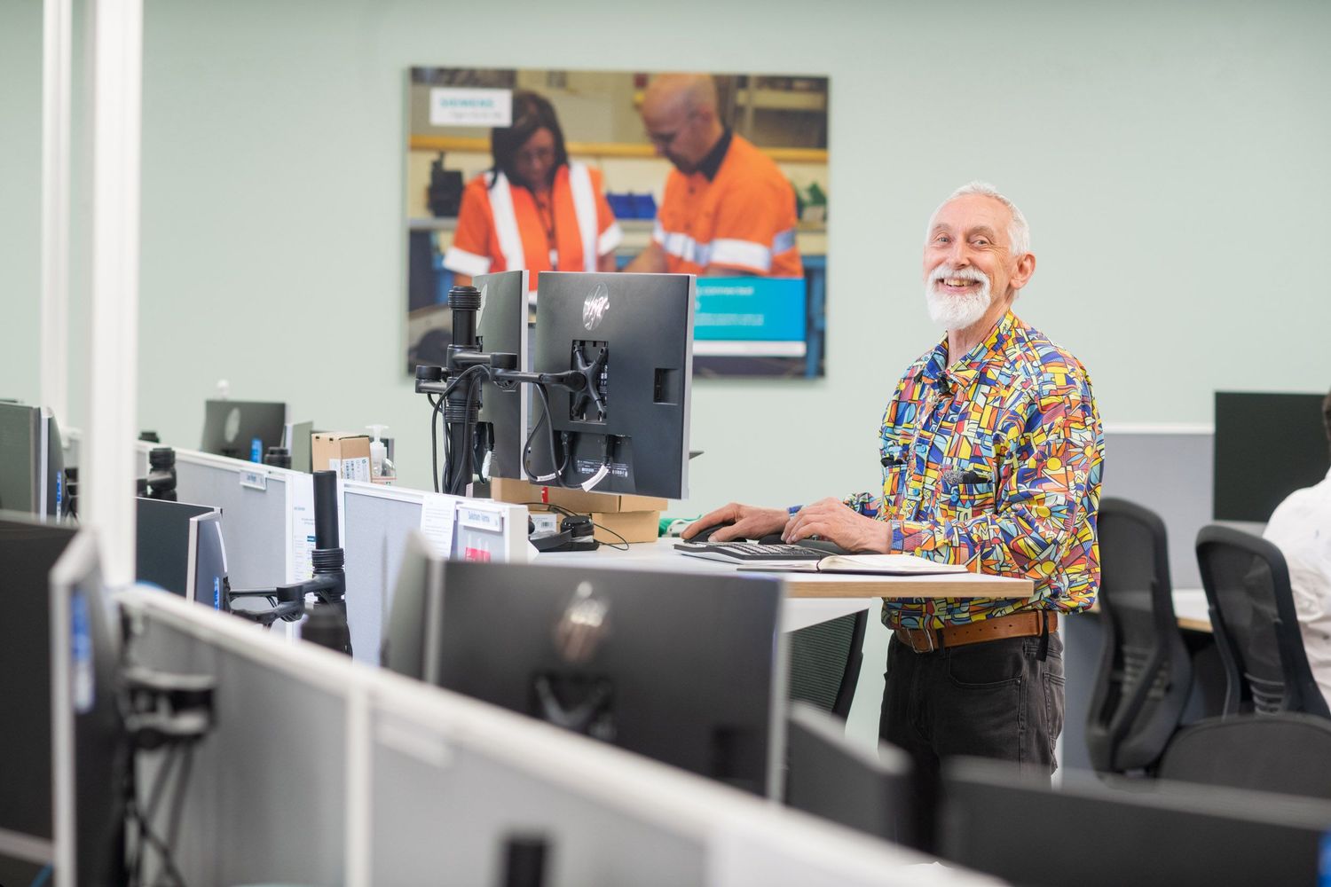 Smiling bearded man in colorful shirt working at standing desk in modern office with monitors.