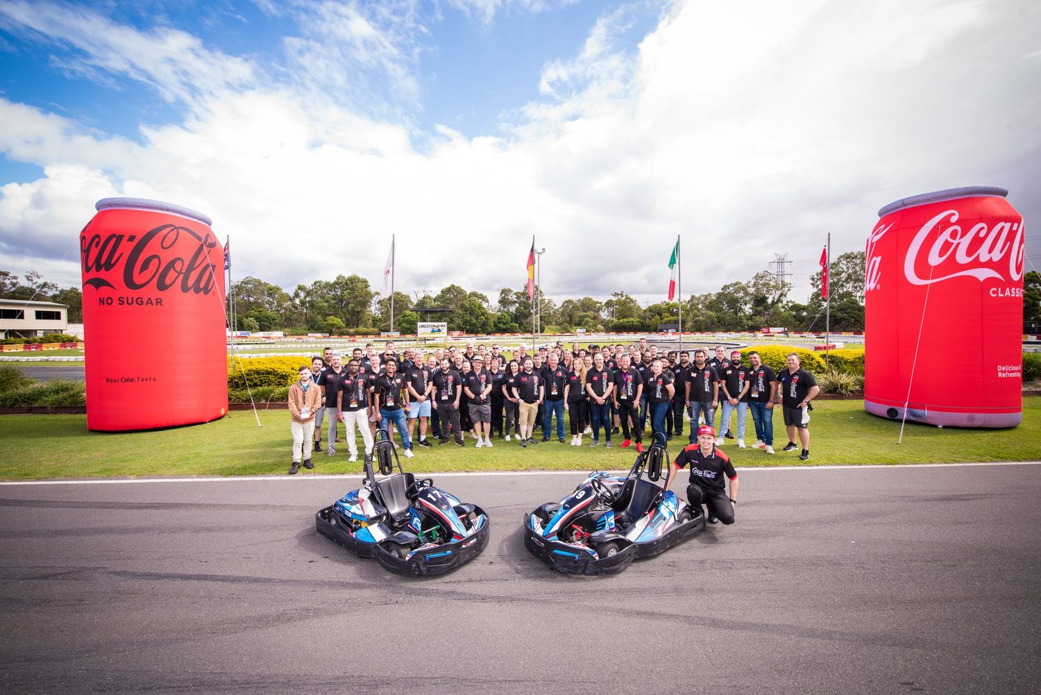 Large corporate group photo at Coca-Cola event with go-karts and giant inflatable Coke cans outdoors.
