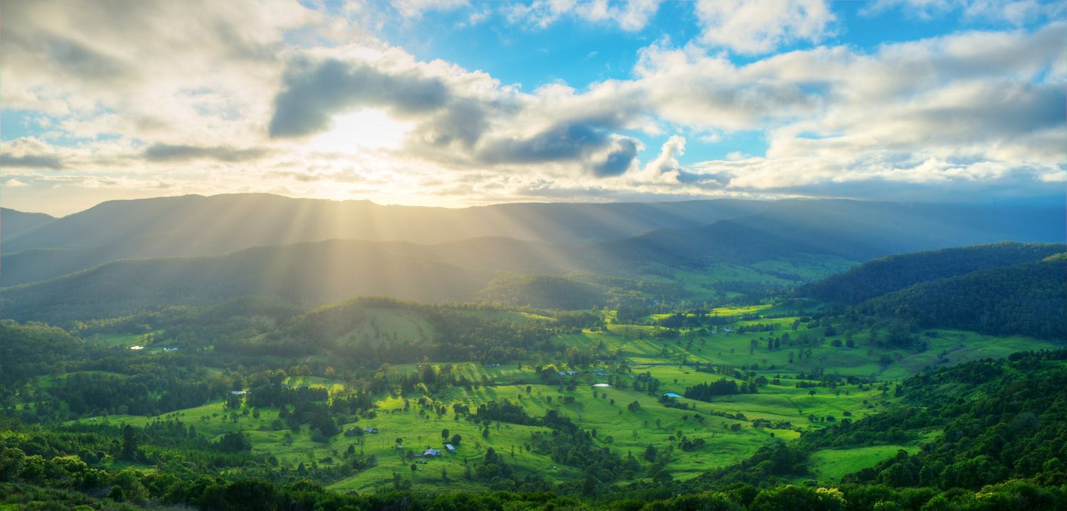 Dramatic sunrays burst through clouds over a lush green valley surrounded by rolling mountains at sunset.
