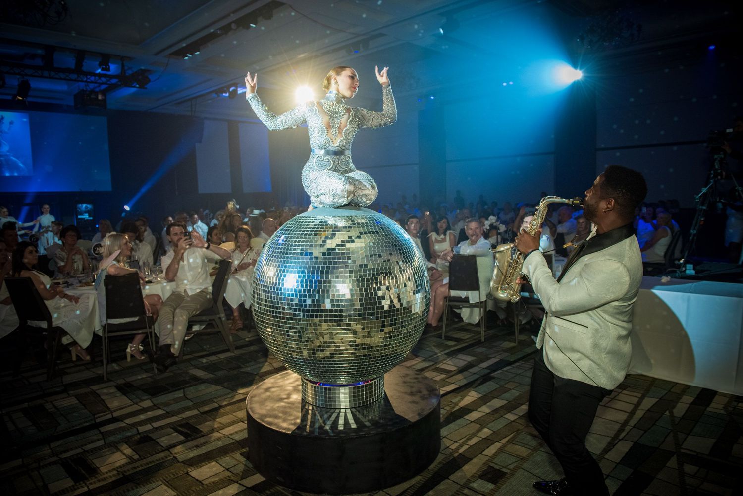 Spectacular performer atop giant disco ball entertained by saxophonist at gala dinner.
