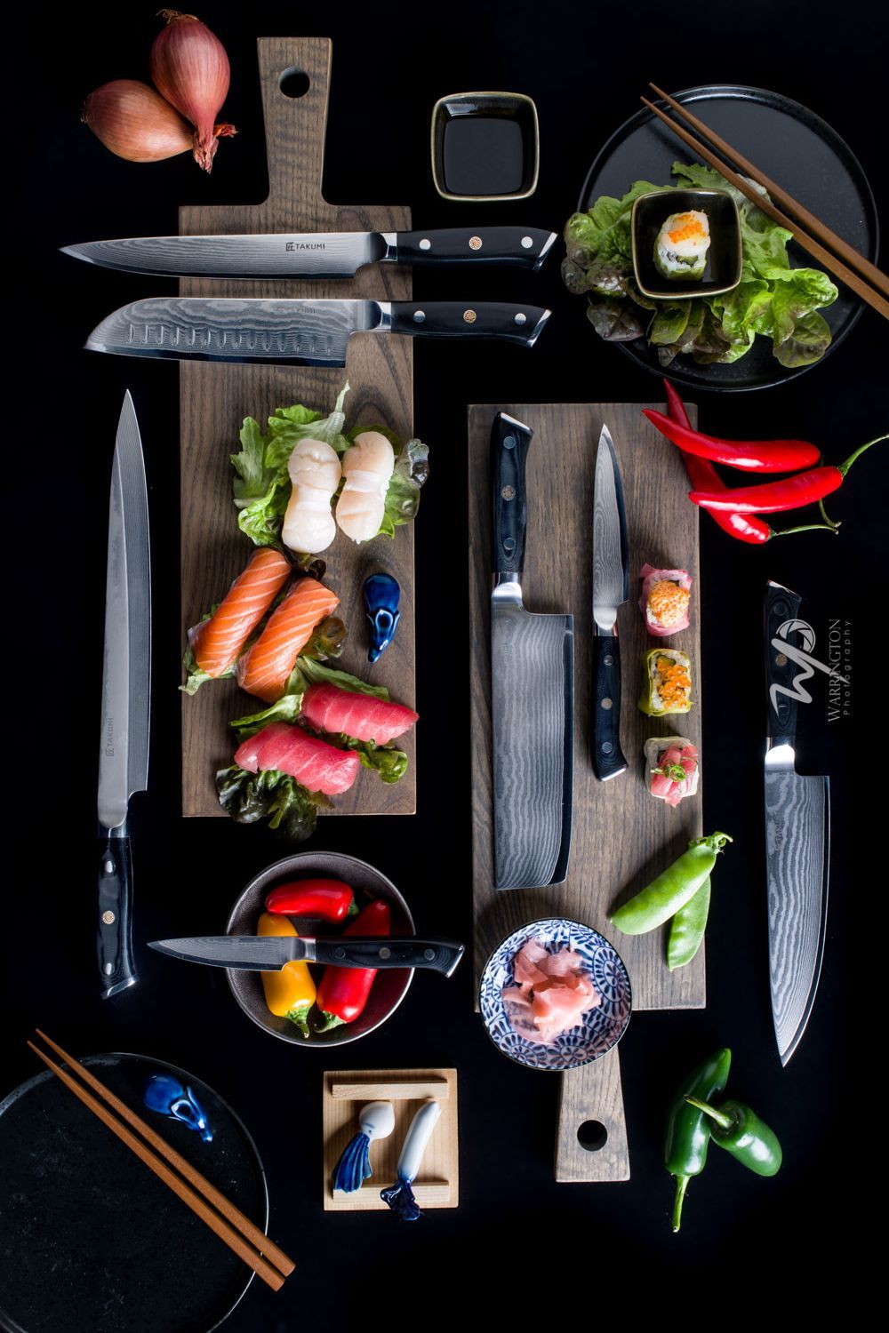 Flat lay of Damascus steel chef knives with fresh sushi ingredients on wooden cutting boards on black background.