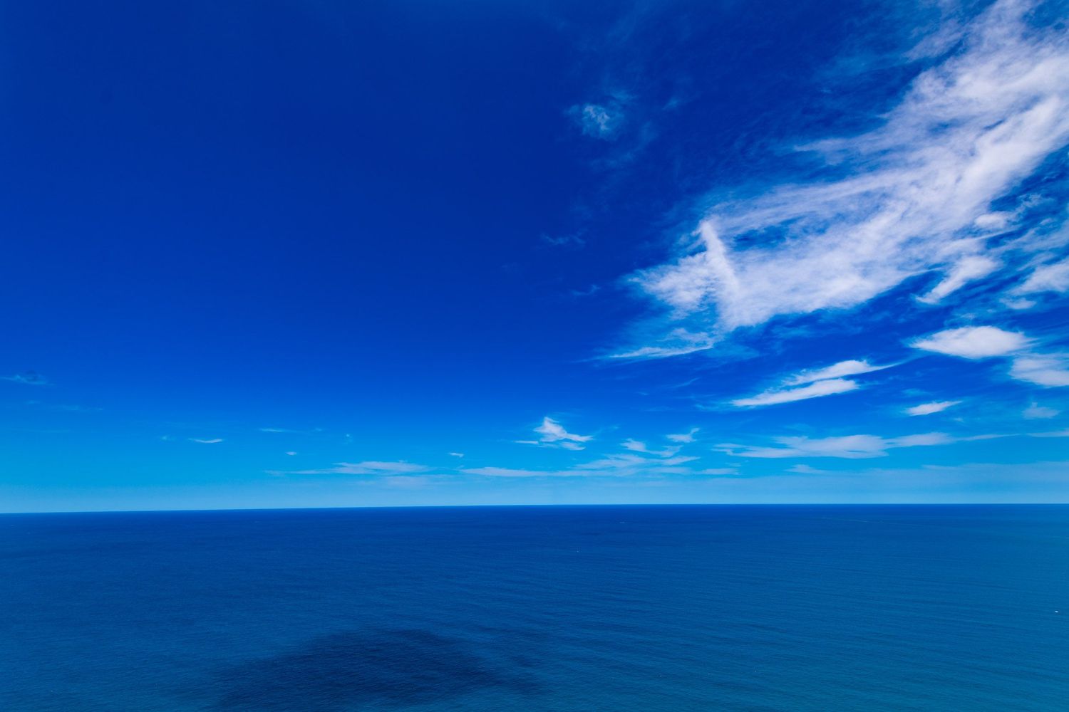 Vibrant deep blue ocean under a clear blue sky with wispy white clouds.