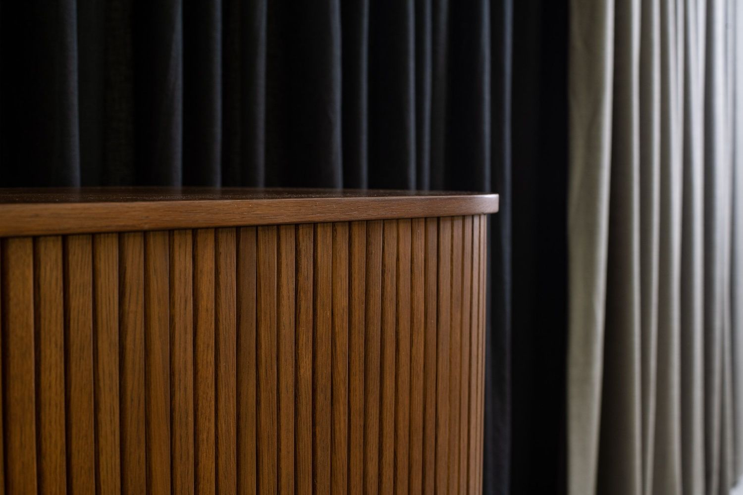 Close-up of a mid-century modern wooden cabinet with vertical slat detailing beside dark curtains in an interior space.