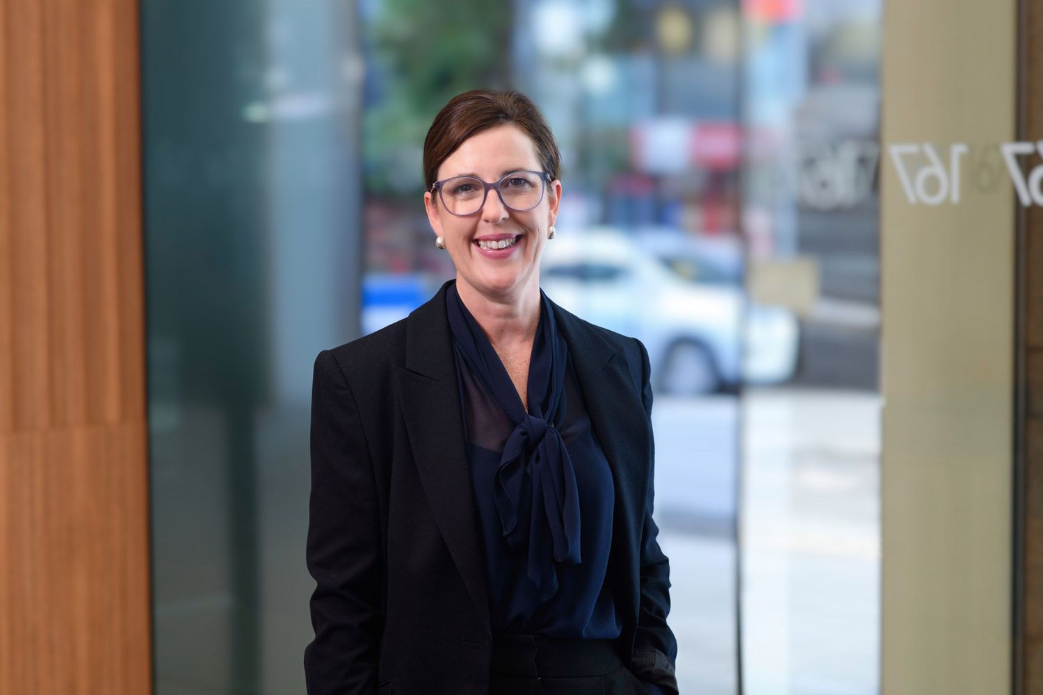 Professional woman in black blazer smiles in modern urban building lobby with city street behind.